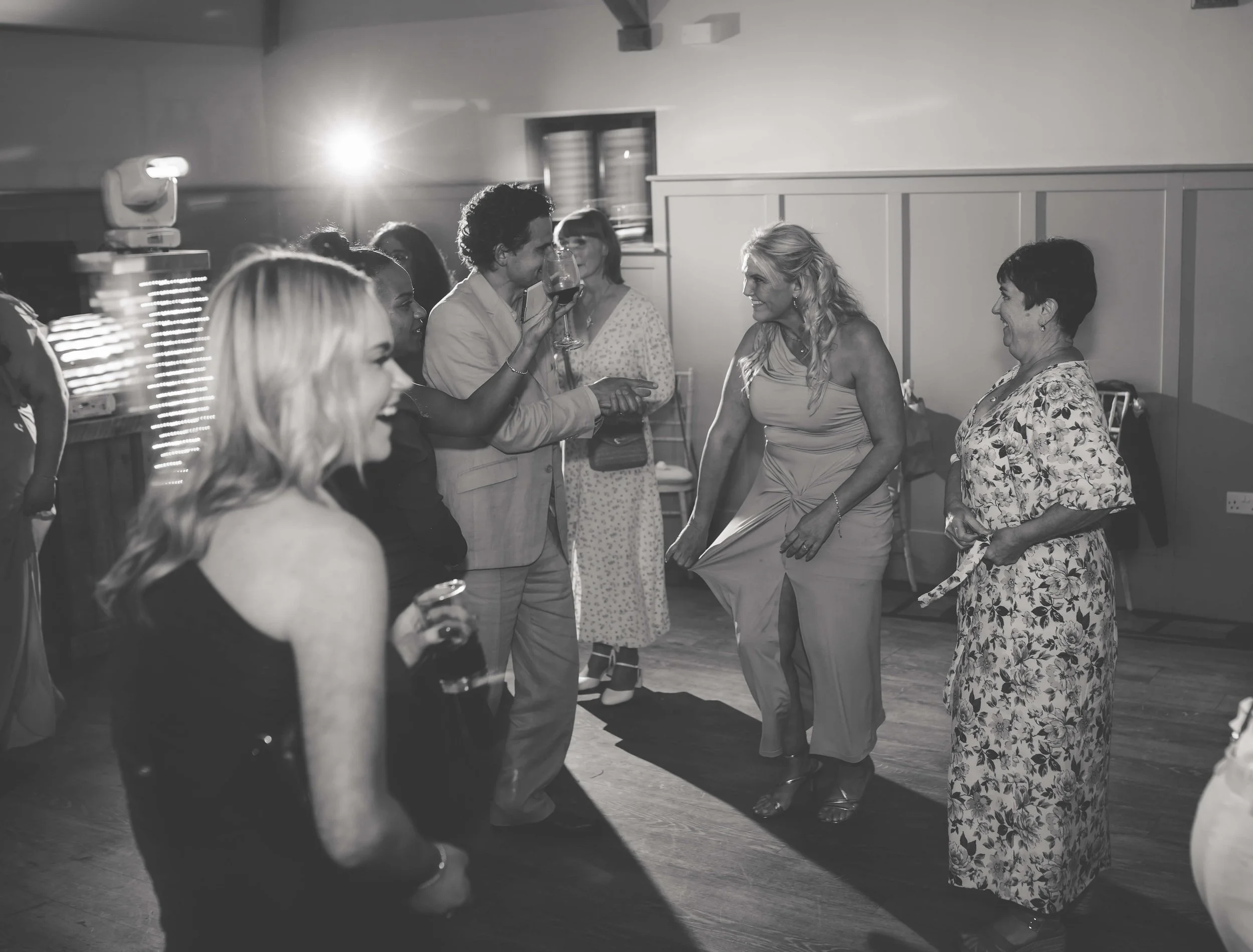 Group of people at a celebration or party, dancing and socializing, with a woman holding a glass of wine and others smiling and chatting in a room with wood flooring.