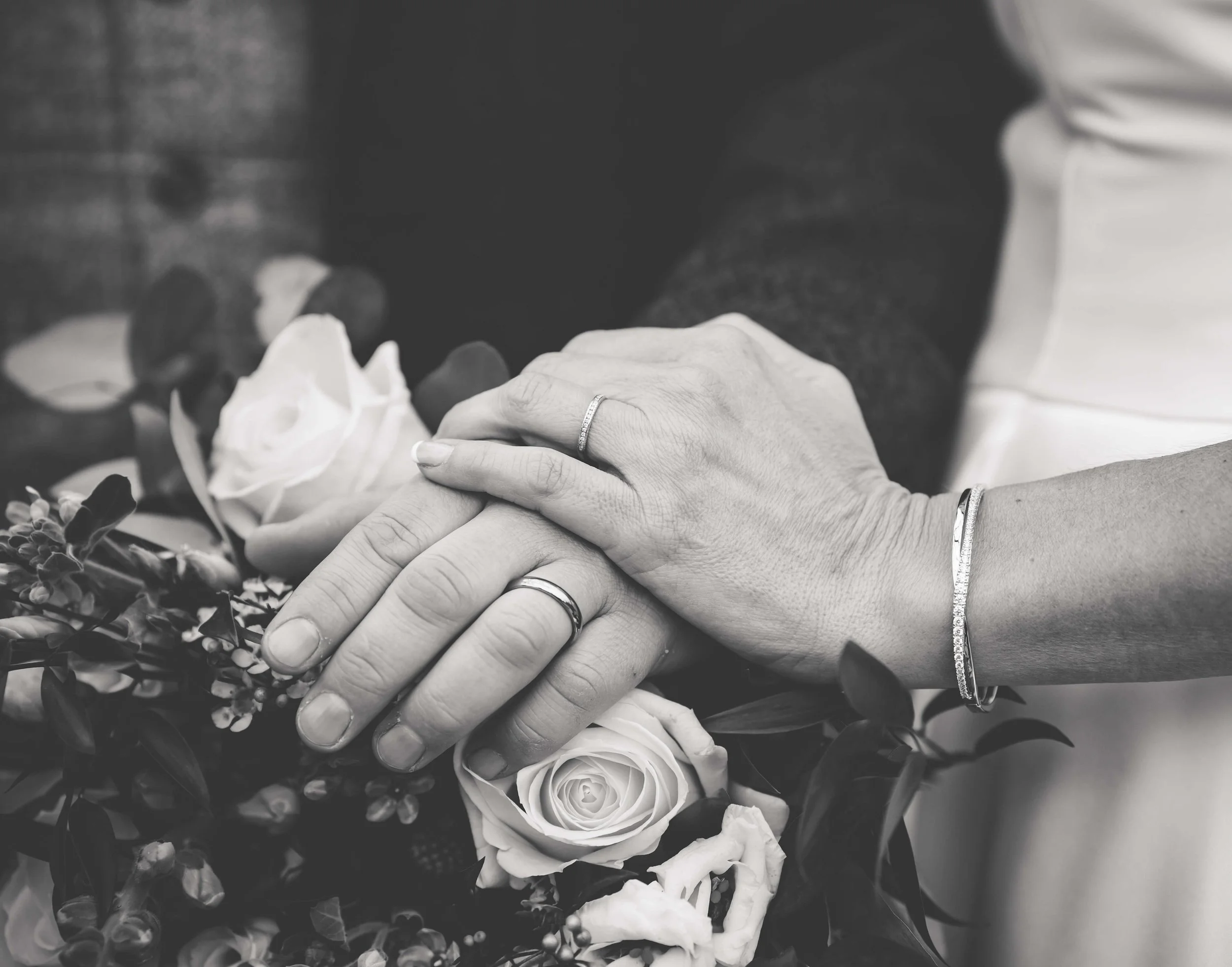 Close-up of a couple's hands with wedding rings, holding a flower bouquet, in black and white.