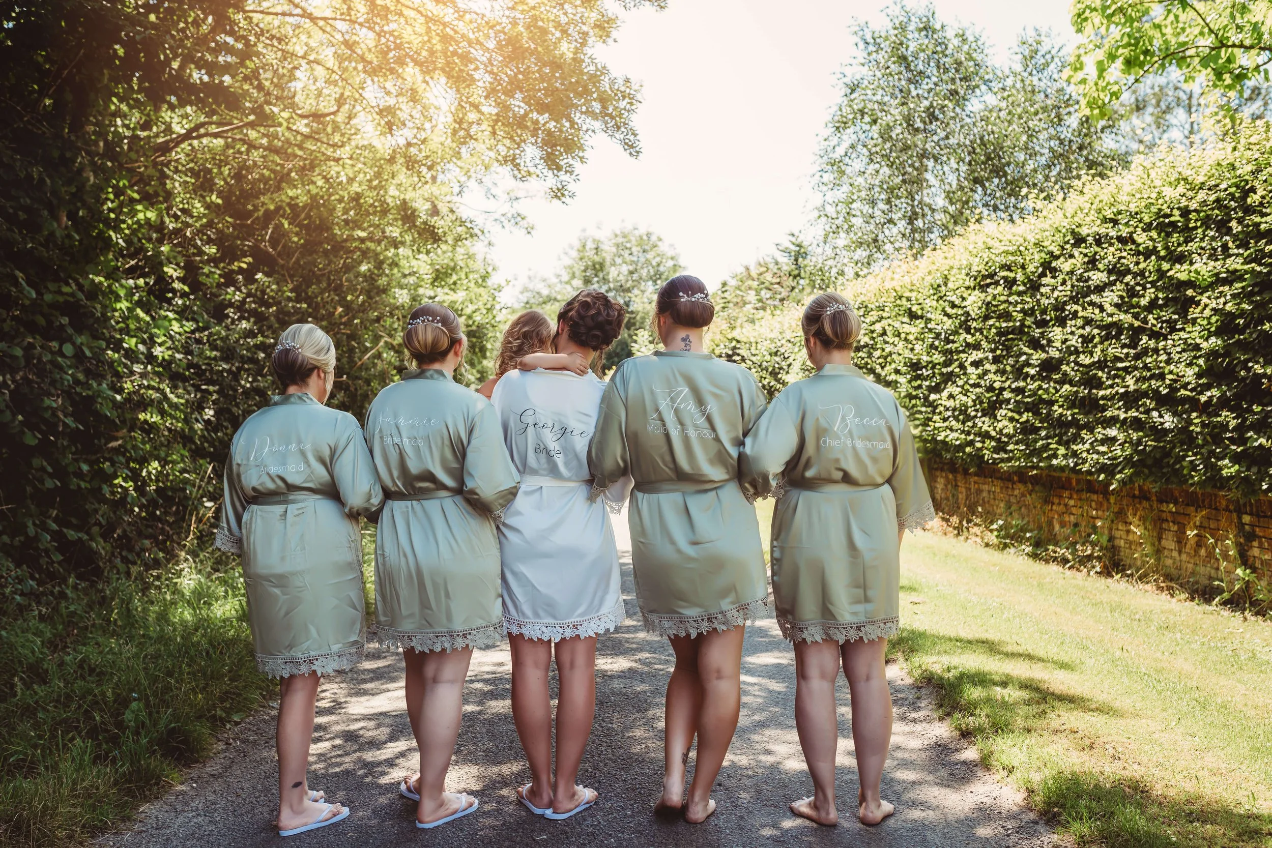 A group of six women, including a bride, walking on a path in a green, sunlit outdoor setting, with four women in matching satin robes with lace trim and the bride in a white robe. The robes have names and roles embroidered on the back, such as 'Brid