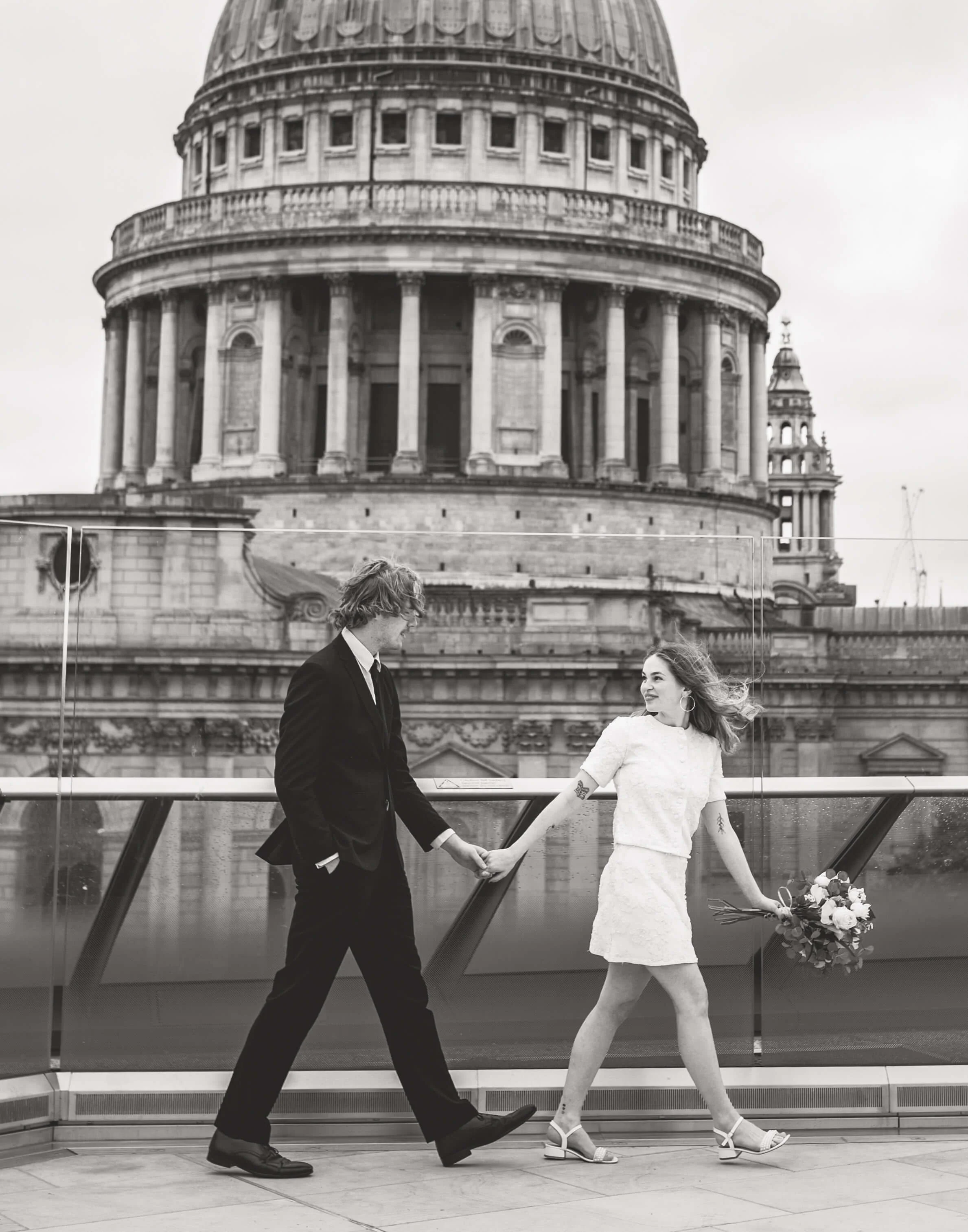 A couple holding hands on a rooftop with St. Paul's Cathedral in London in the background. The woman is wearing a white dress and carrying a bouquet, while the man is in a black suit.