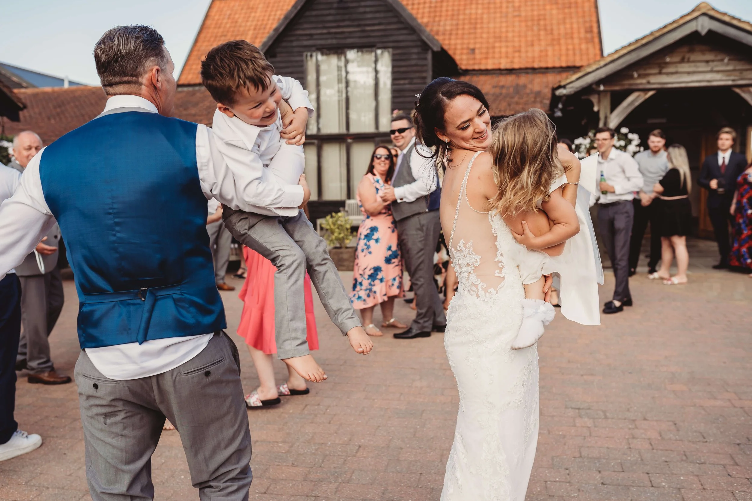 A bride in a wedding dress holding a young girl, with an older man lifting a young boy on his shoulder, at an outdoor wedding reception, with guests in the background.