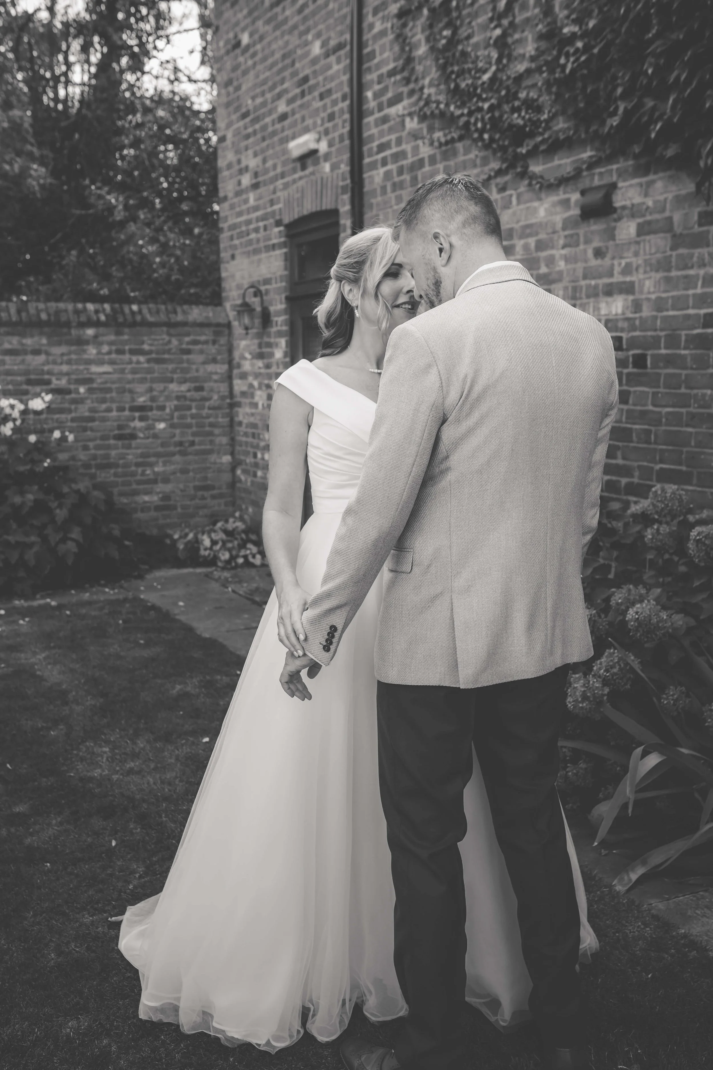 A black and white photo of a bride and groom holding hands, standing close together outside near a brick wall, sharing an intimate moment.