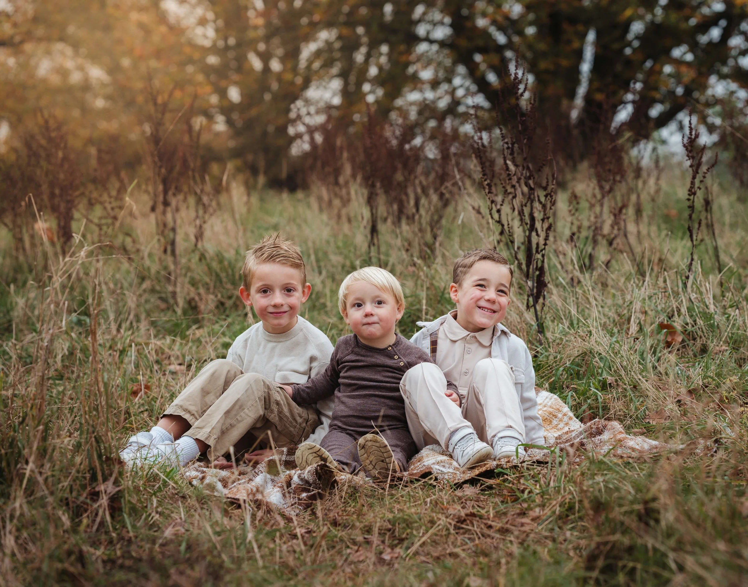 Three young boys sitting together on a blanket in a grassy field during fall.