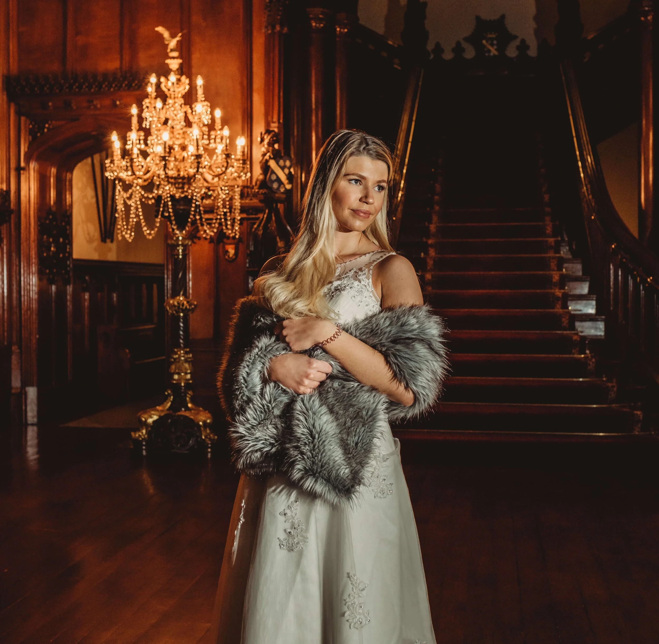 A young woman in a white dress holding a fur stole stands on a grand staircase in an elegant, wood-paneled room with a chandelier.