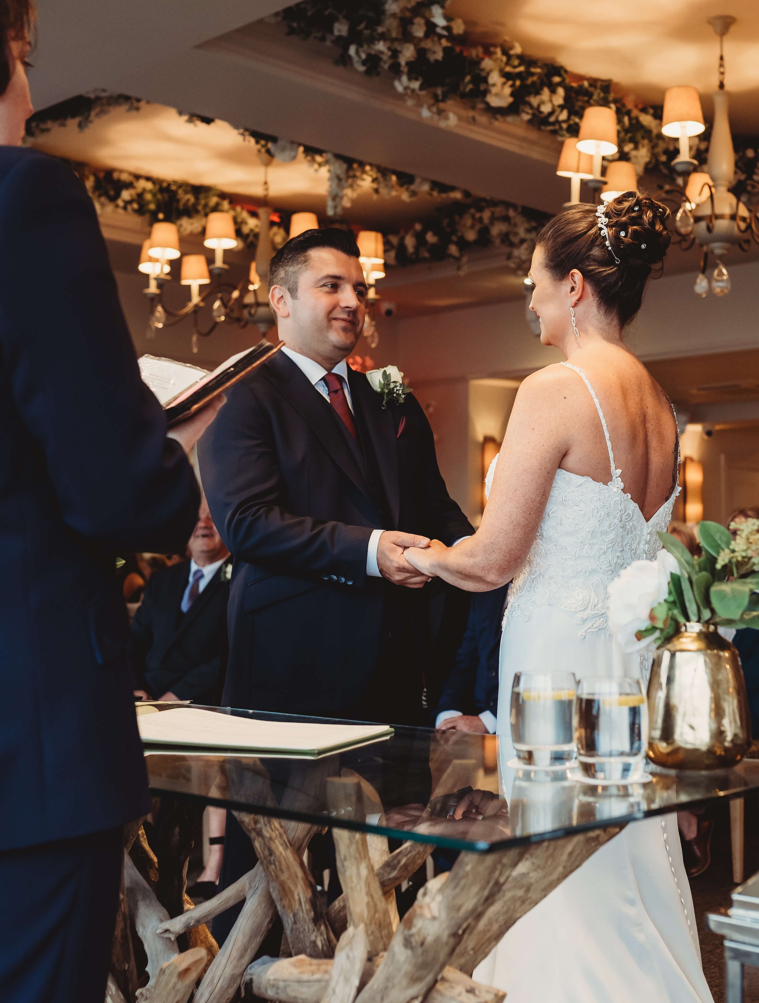 A bride and groom holding hands and exchanging vows during a wedding ceremony, with the officiant standing to the side, in a decorated indoor venue.