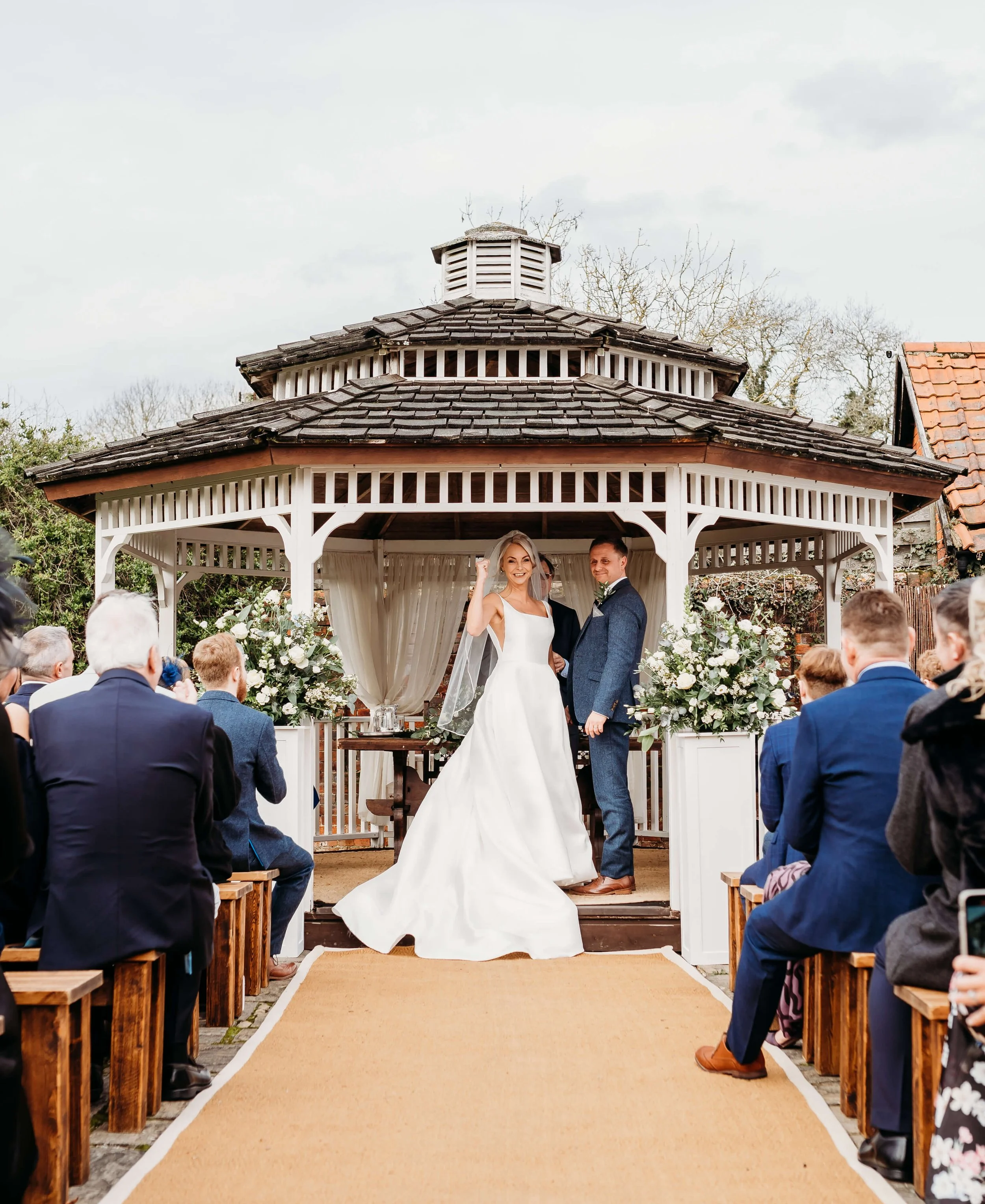 Wedding ceremony outdoors at a gazebo with the bride and groom standing in front of guests, with floral decorations and a beige carpet on the ground.