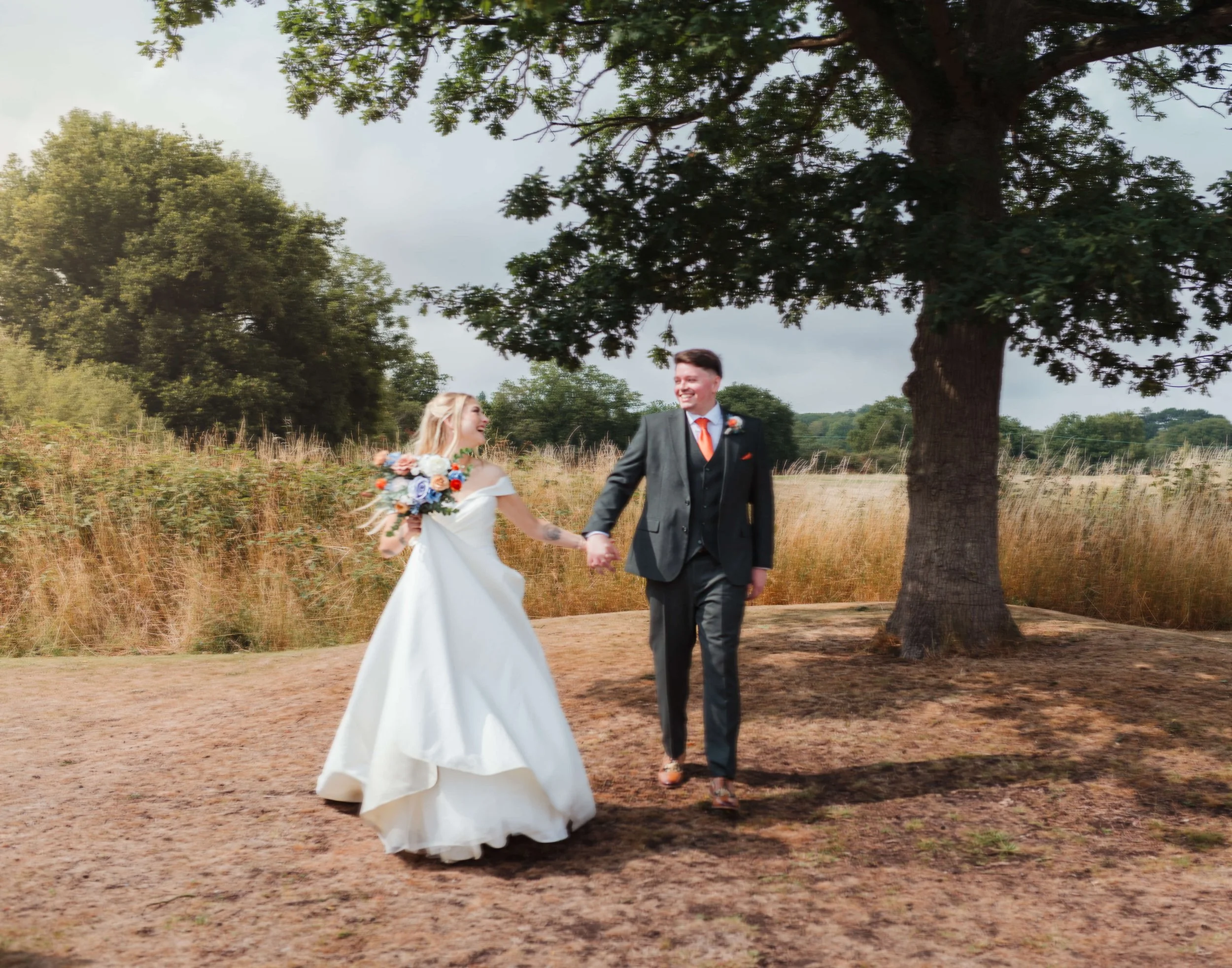 A bride and groom holding hands and walking outdoors beside a large tree, with grassy fields and a partly cloudy sky in the background.