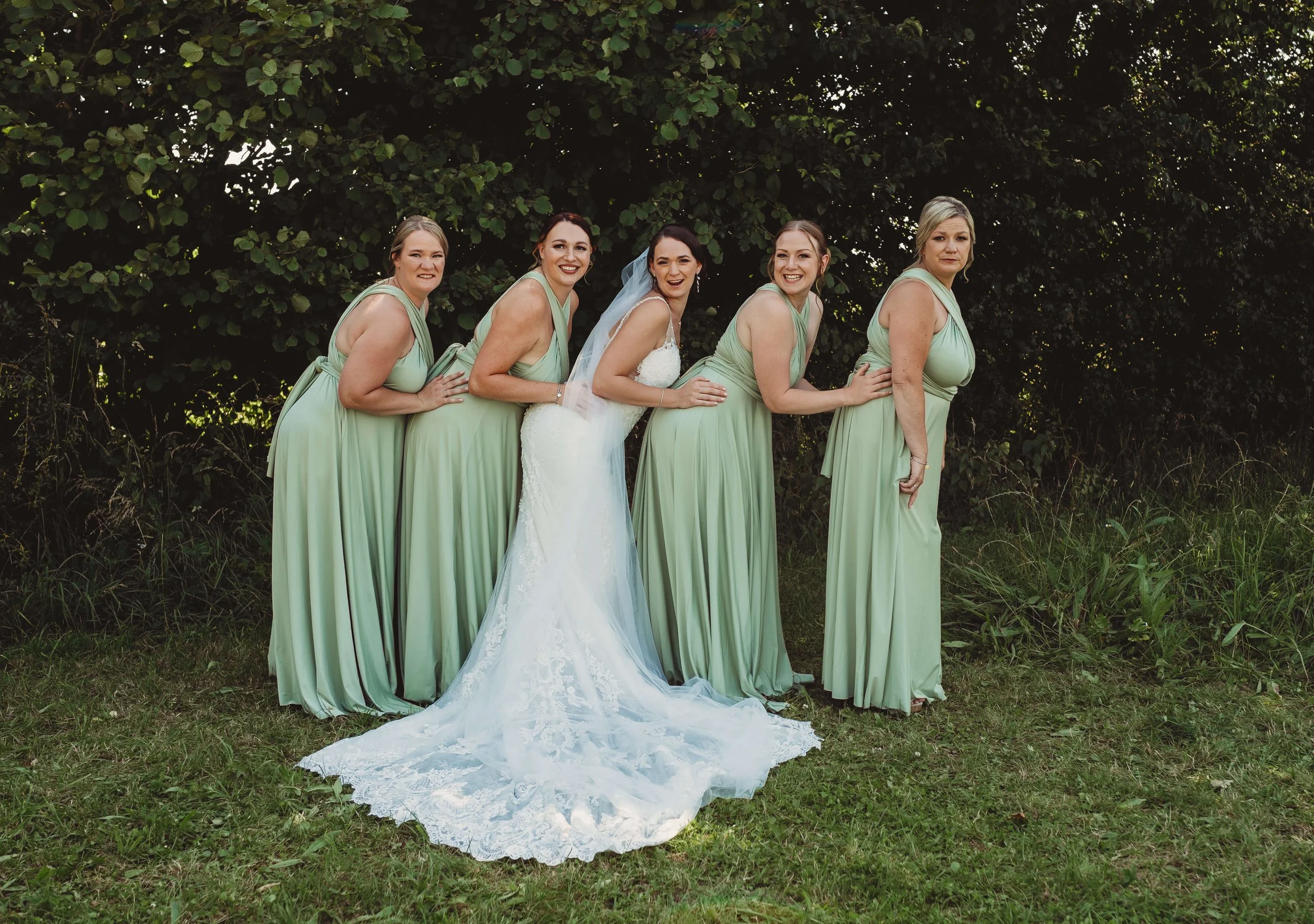 Bride in a white wedding gown with lace train posing outdoors with five bridesmaids dressed in matching light green dresses, all standing in front of green foliage and grass.