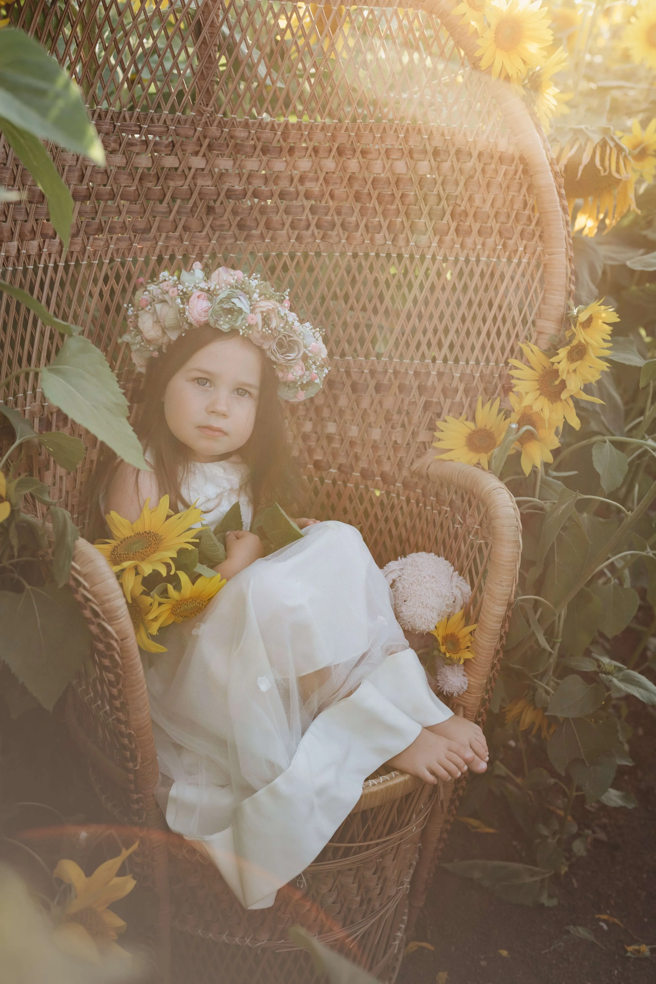 A young girl with a floral crown sitting on a wicker chair surrounded by sunflowers.