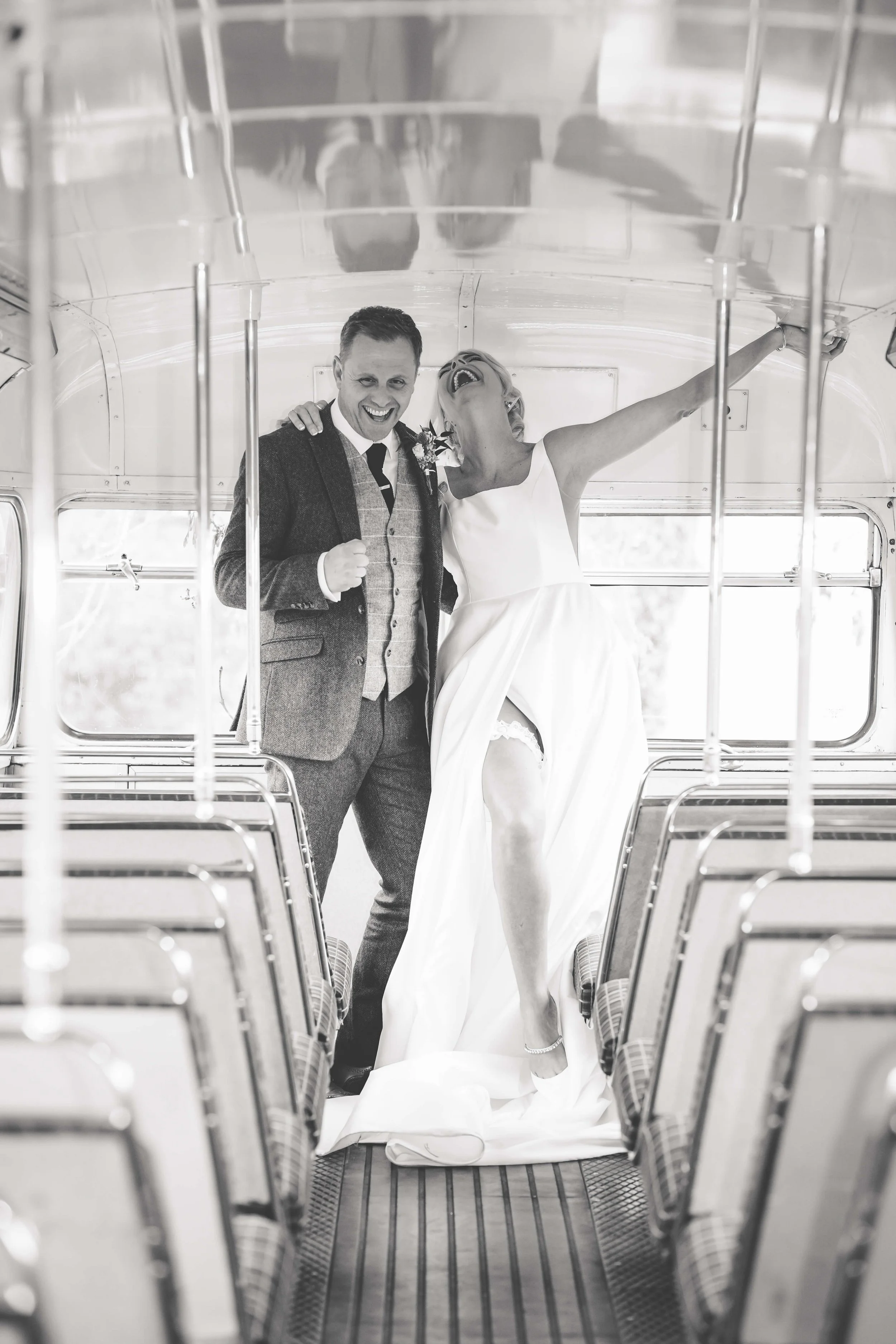 Black and white photo of a bride and groom celebrating inside a bus, with the bride lifting her leg and both smiling.