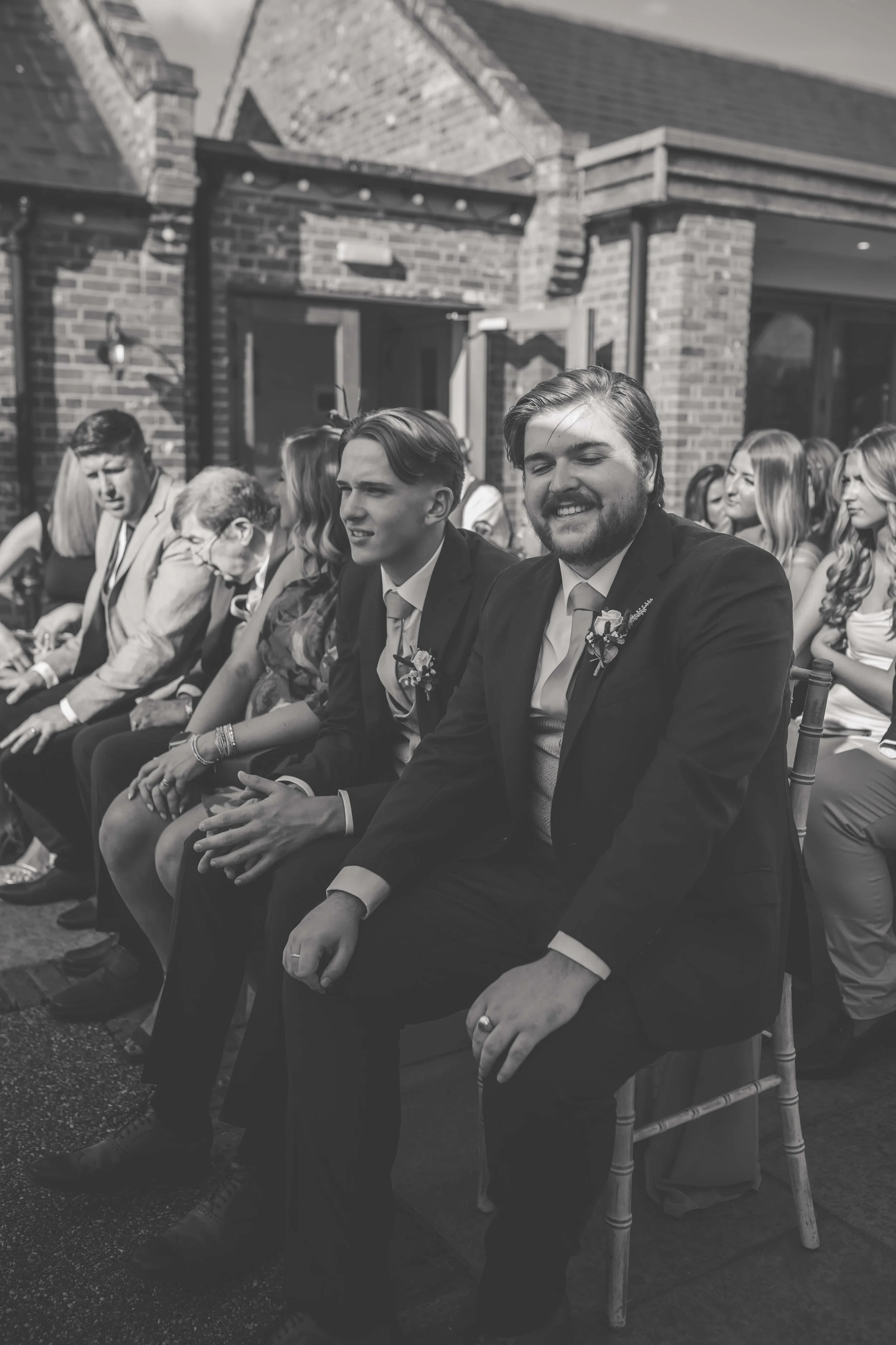 A group of people seated outdoors at a wedding or formal event, with the two men in the foreground wearing suits and boutonnieres, smiling and enjoying the occasion. Behind them are women and men also dressed formally, with a brick building in the ba