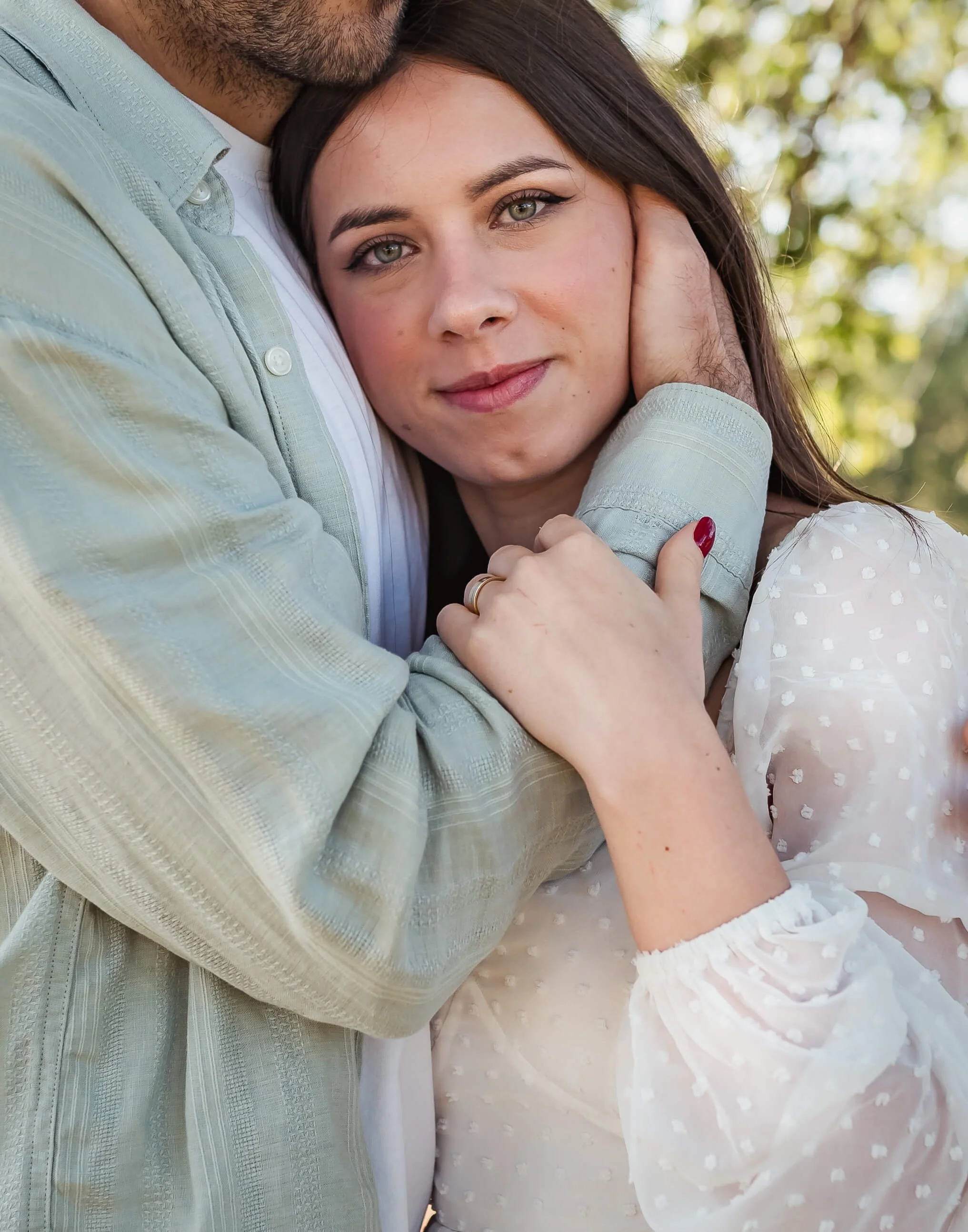 A woman with long dark hair and bright eyes being embraced by a man. She is looking at the camera with a gentle smile, wearing a white dress with sheer dotted fabric. The man, wearing a light-colored shirt, is hugging her tightly, with only part of h