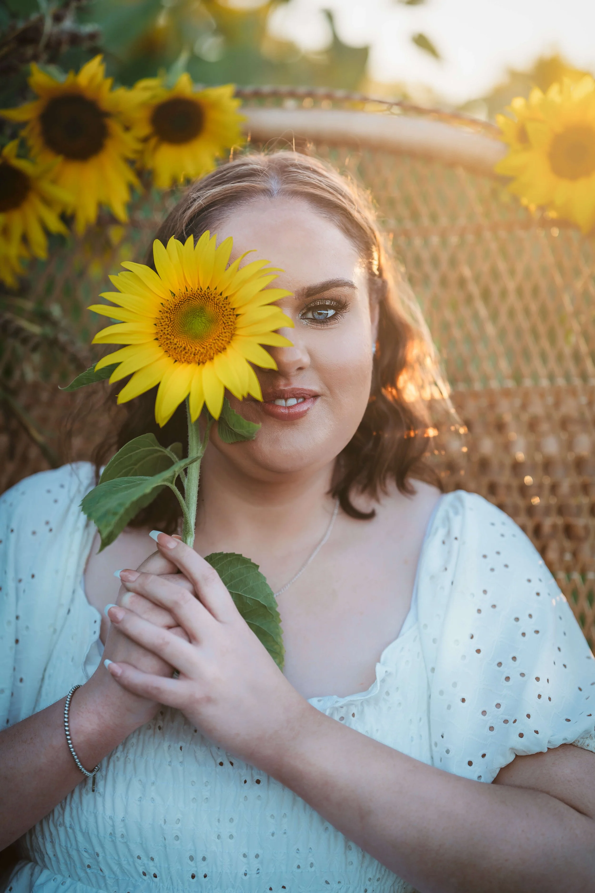 A woman holding a sunflower close to her face, partially covering one eye, standing outdoors in a sunlit area with a natural background.