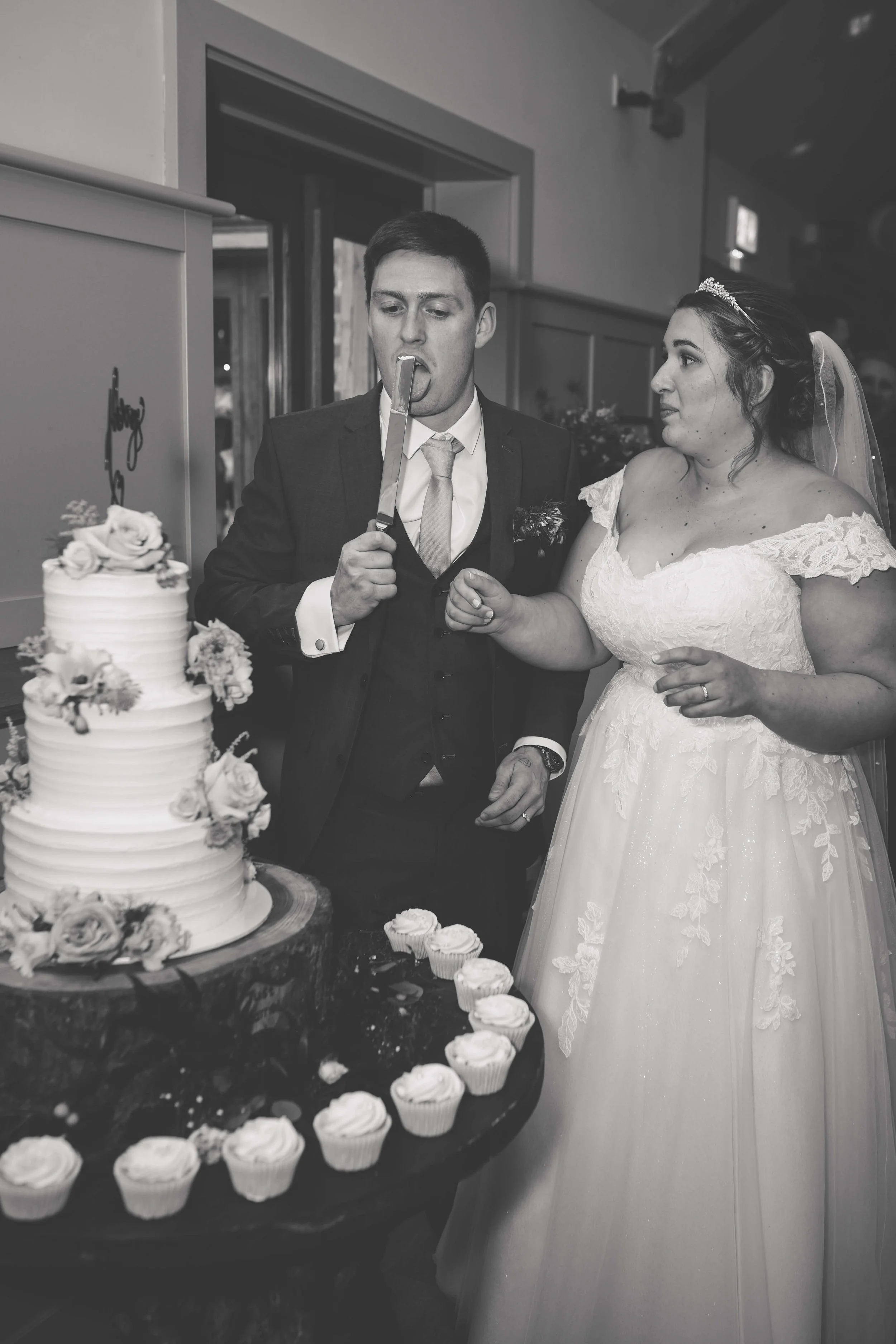 Black and white photo of a bride and groom at their wedding cake cutting, with the groom playfully holding a knife in his mouth.