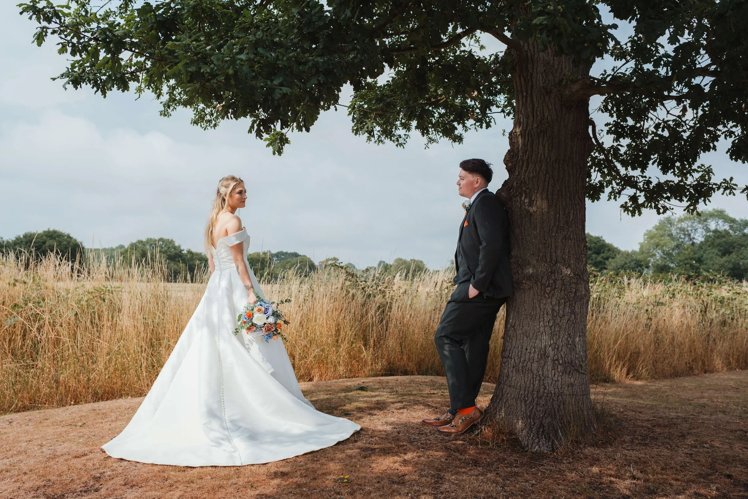 A bride in a white wedding gown holding a bouquet looks at a groom in a black suit leaning against a tree in an outdoor setting with tall grass and a cloudy sky.