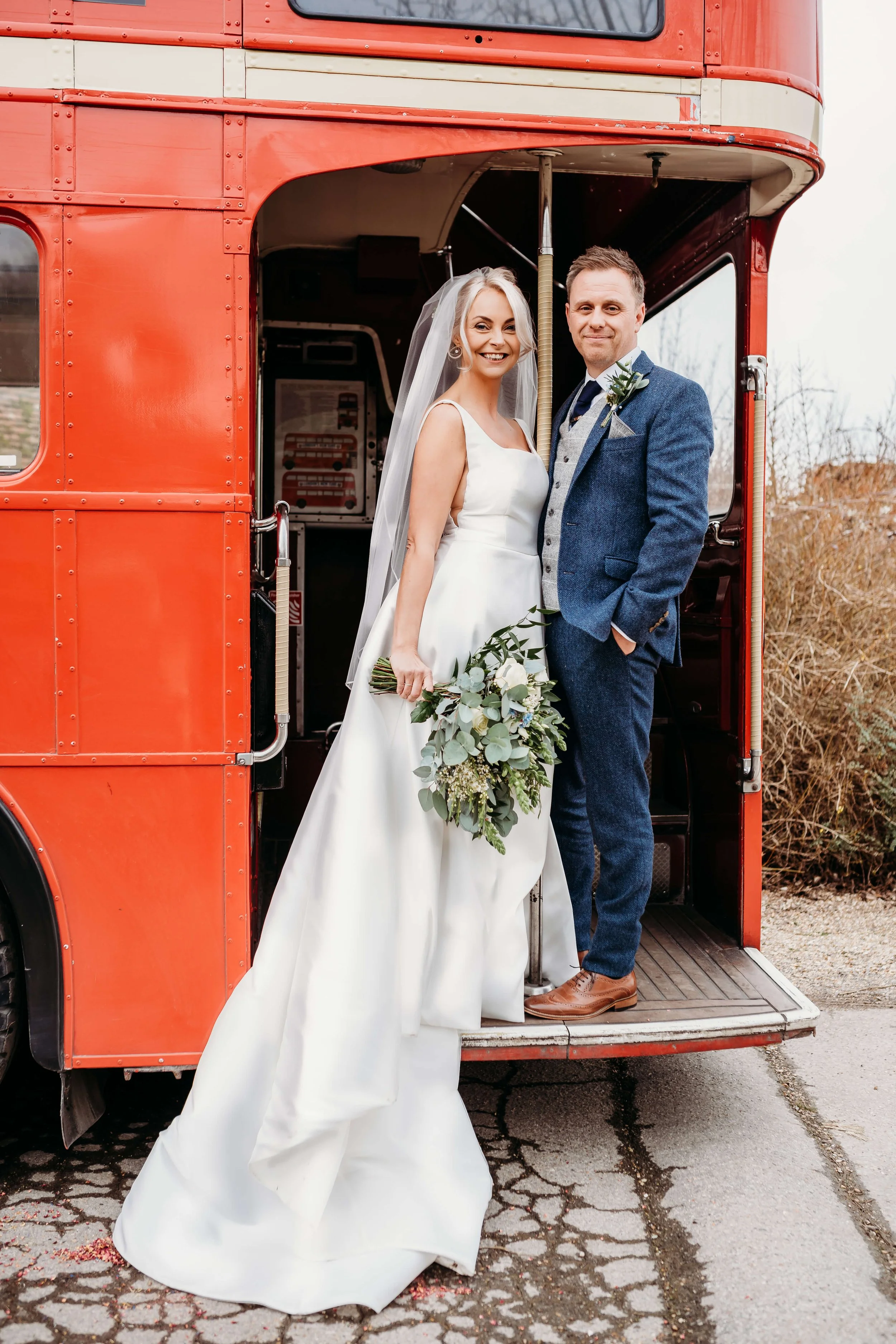 A bride and groom stand inside a vintage red bus on their wedding day, smiling at the camera. The bride is holding a bouquet of greenery and white flowers, wearing a white wedding gown and veil. The groom is dressed in a blue suit with a vest and tie
