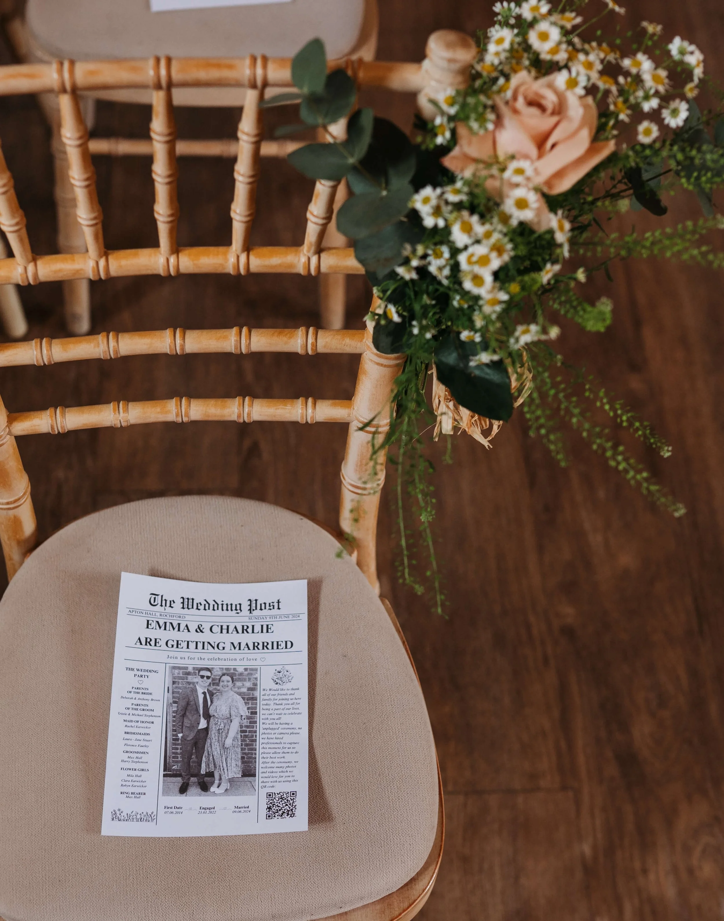A wedding program on a beige chair with a wooden floral decoration attached to the chair back, featuring a bouquet of light pink roses, small white daisies, and greenery.