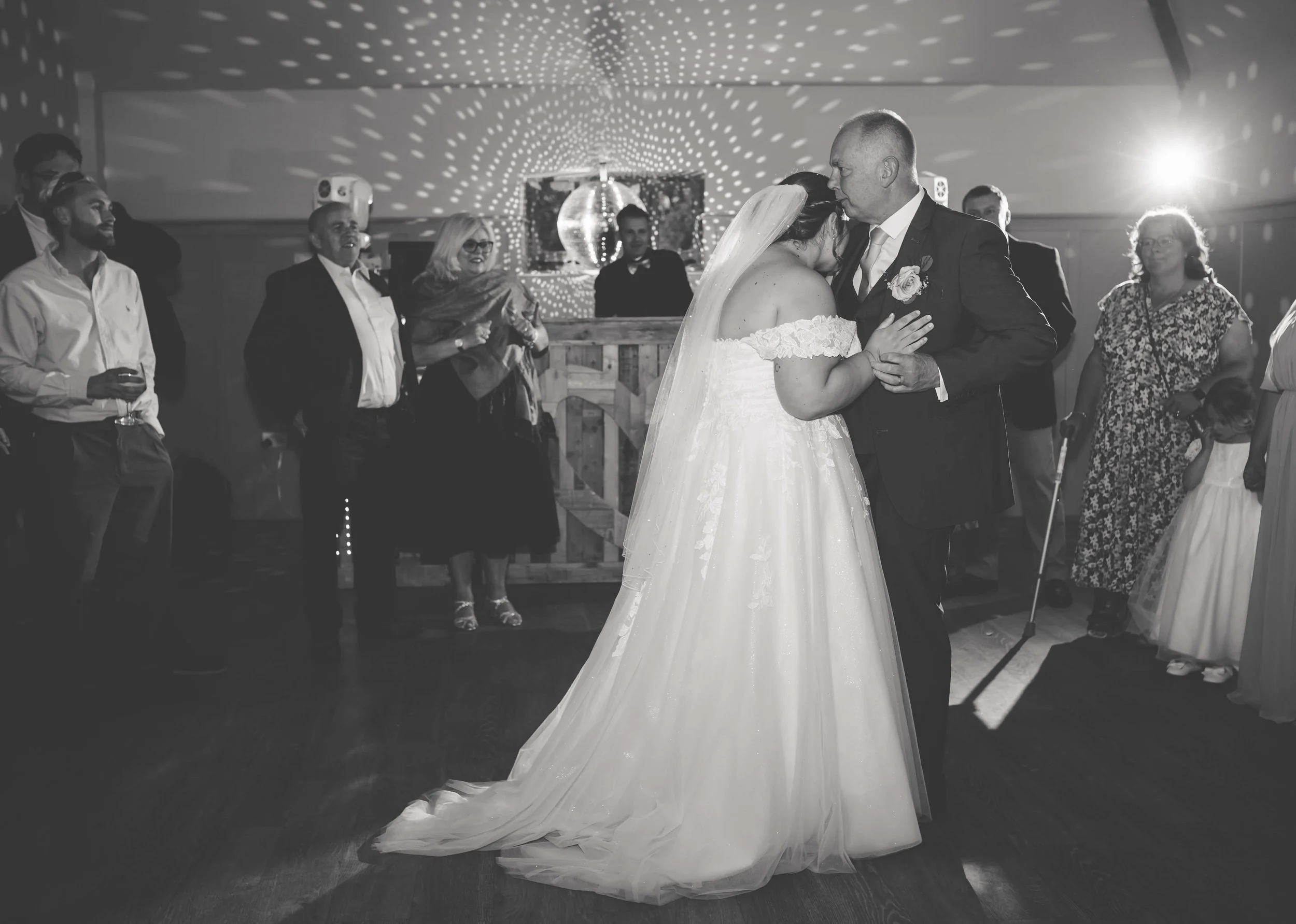 A bride and groom share their first dance at a wedding reception, surrounded by friends and family, with a disco ball and decorative lighting in the background.