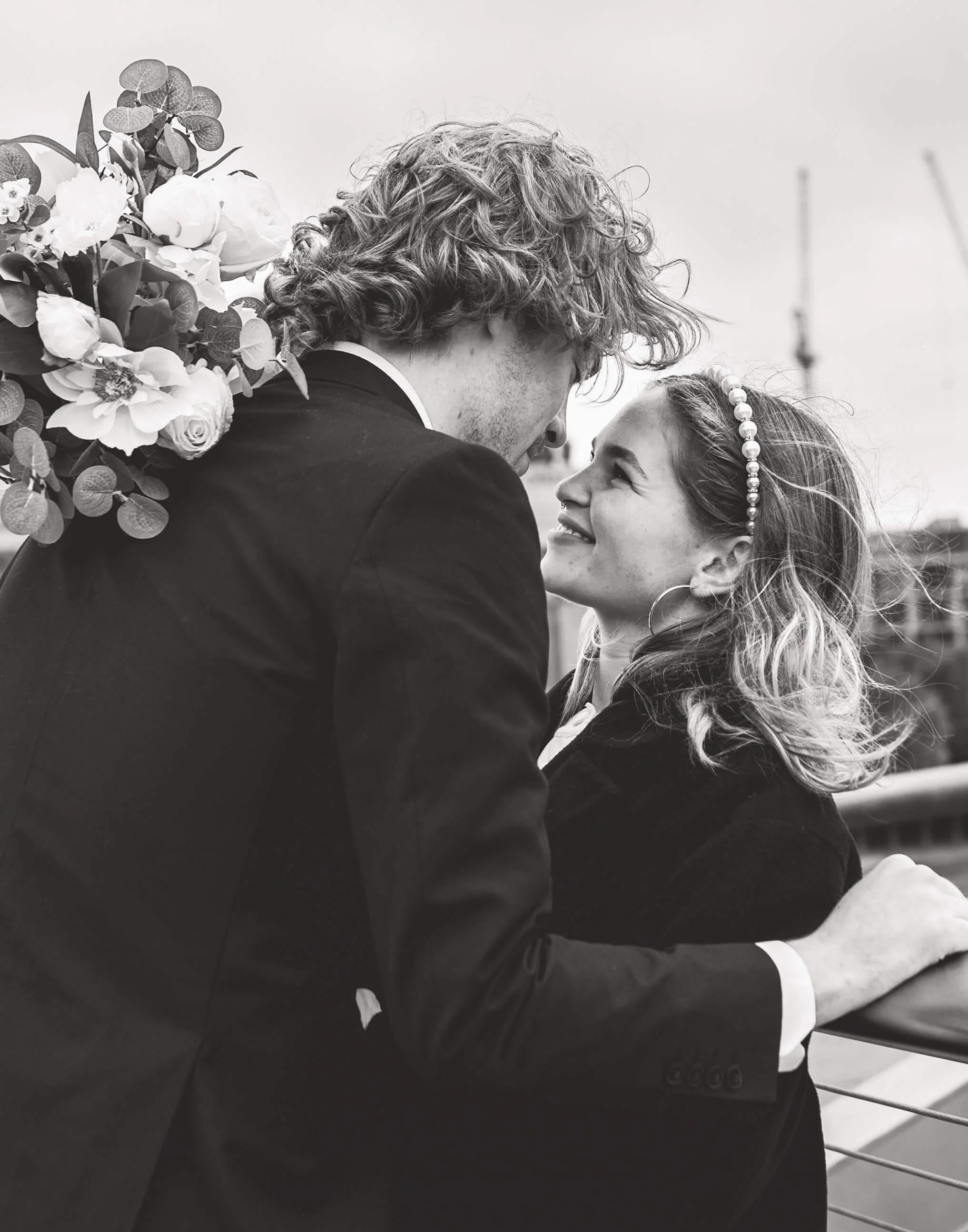 A black and white photograph of a couple at a romantic moment, leaning close to each other, smiling, with the woman wearing a pearl headband and hoop earrings, and the man holding a bouquet of flowers on his shoulder, against a blurred outdoor citysc