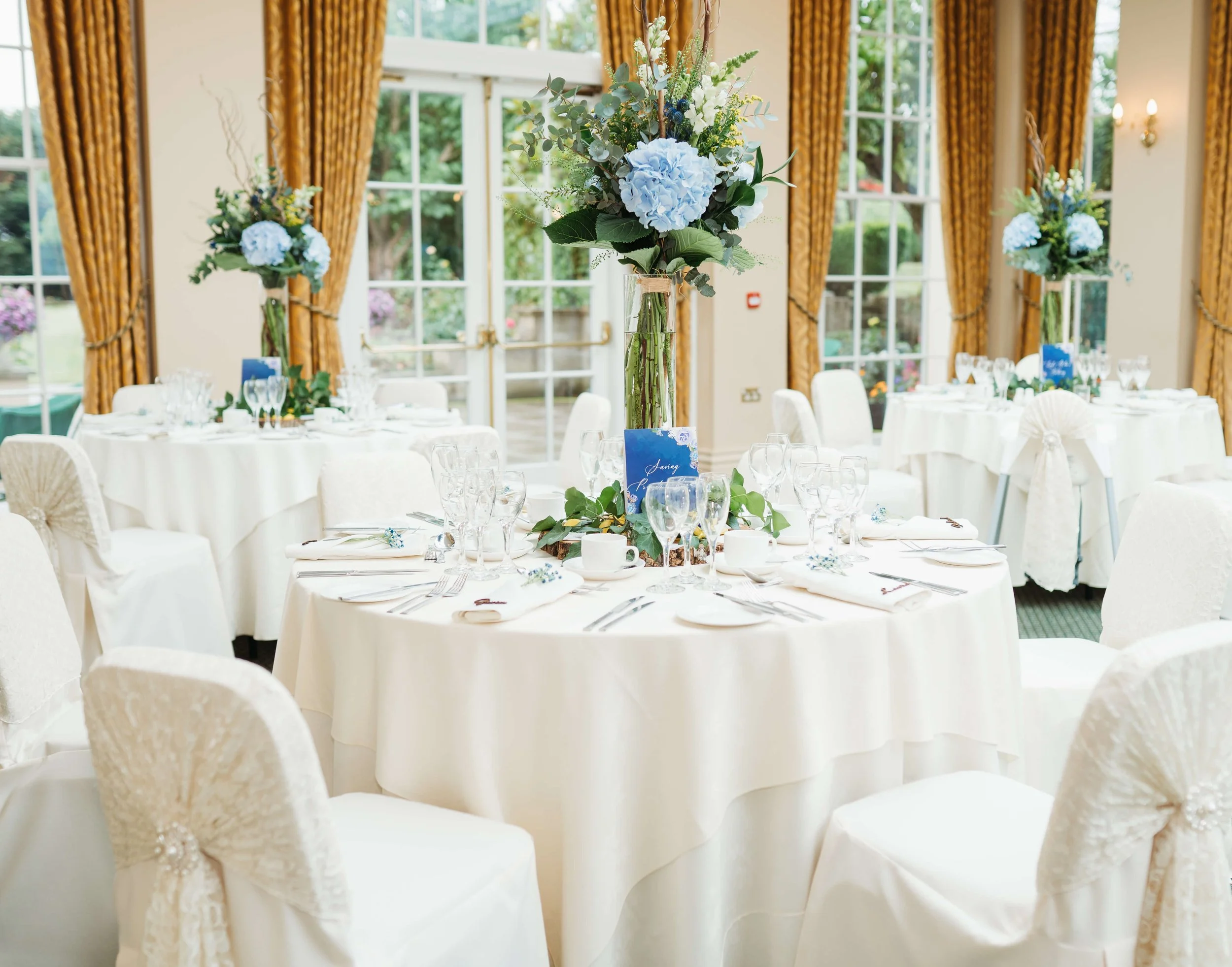 Wedding banquet hall with round tables covered in white tablecloths, decorated with blue floral centerpieces in tall glass vases, surrounded by white chairs with fabric covers and bows. Large windows with gold curtains reveal a garden outside.