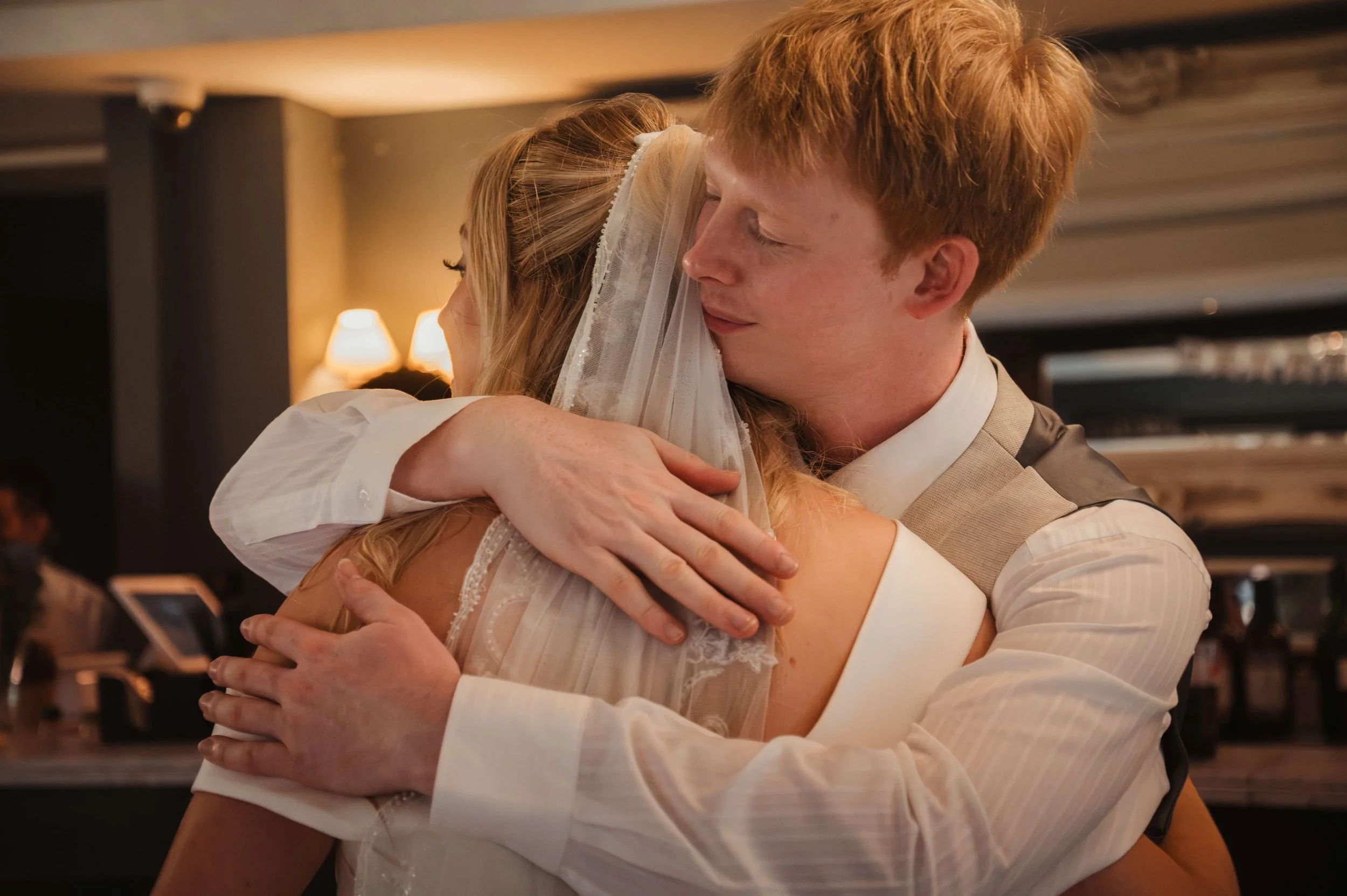 A man and woman embracing tightly at a wedding reception, with warm lighting and a blurry background.