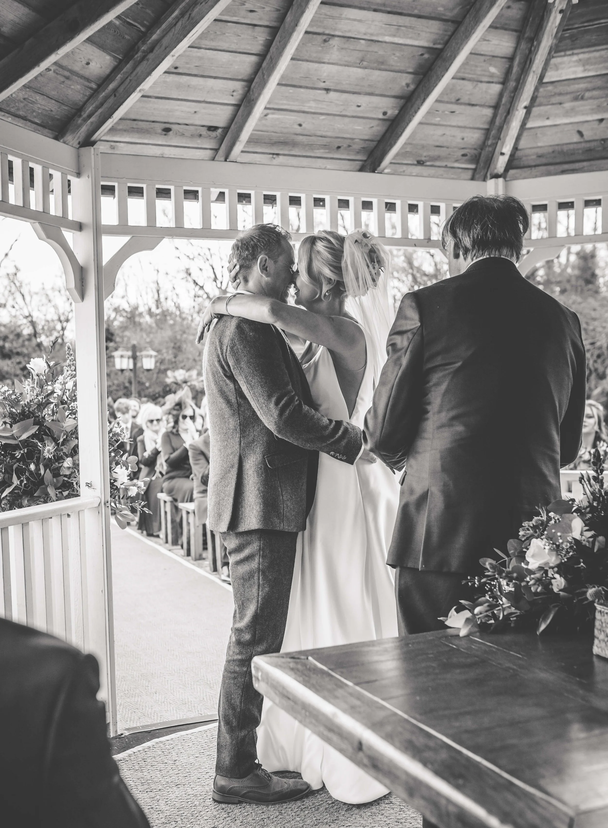 A bride and groom embrace during their wedding ceremony under a gazebo, with guests seated in the background.