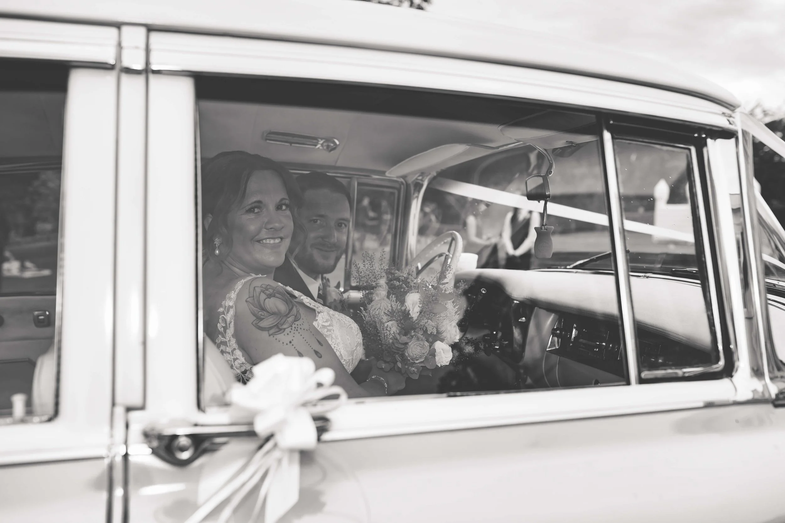 A black and white photo of a bride and groom sitting inside a vintage car, smiling at the camera. The bride holds a bouquet of flowers, and the car is decorated with ribbons.
