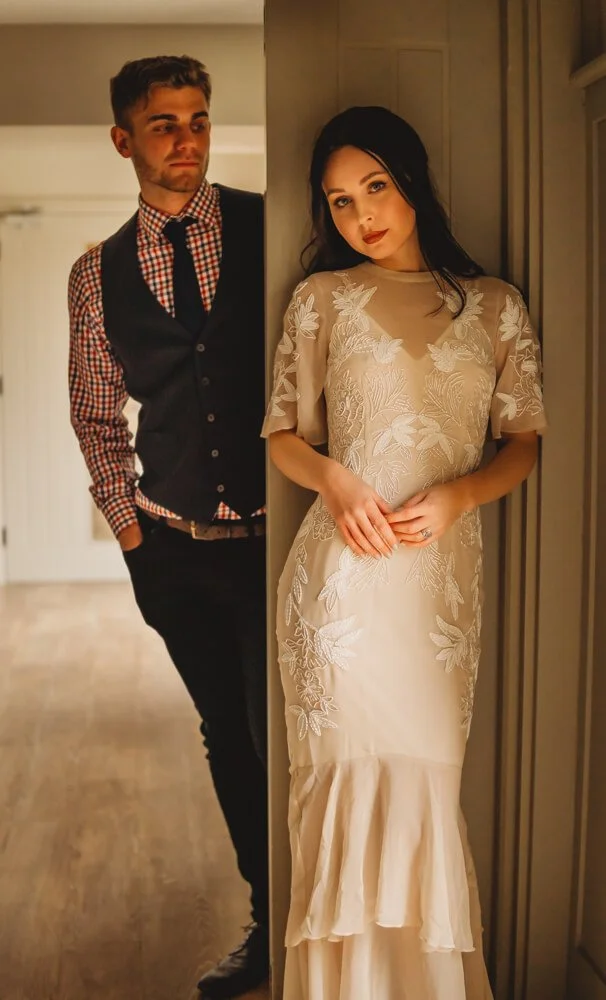A young man and woman stand near a wall indoors, the man peering at the woman, who is wearing a beige dress with floral embroidery, as she looks thoughtfully ahead.