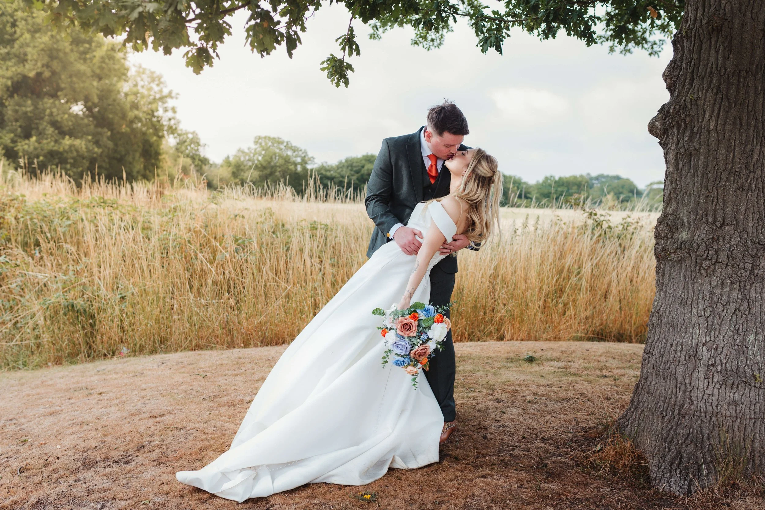 A newlywed couple shares a romantic kiss outdoors under a tree, the groom in a black suit with an orange tie, and the bride in a white wedding dress holding a colorful bouquet, in a natural setting with tall grass and trees.