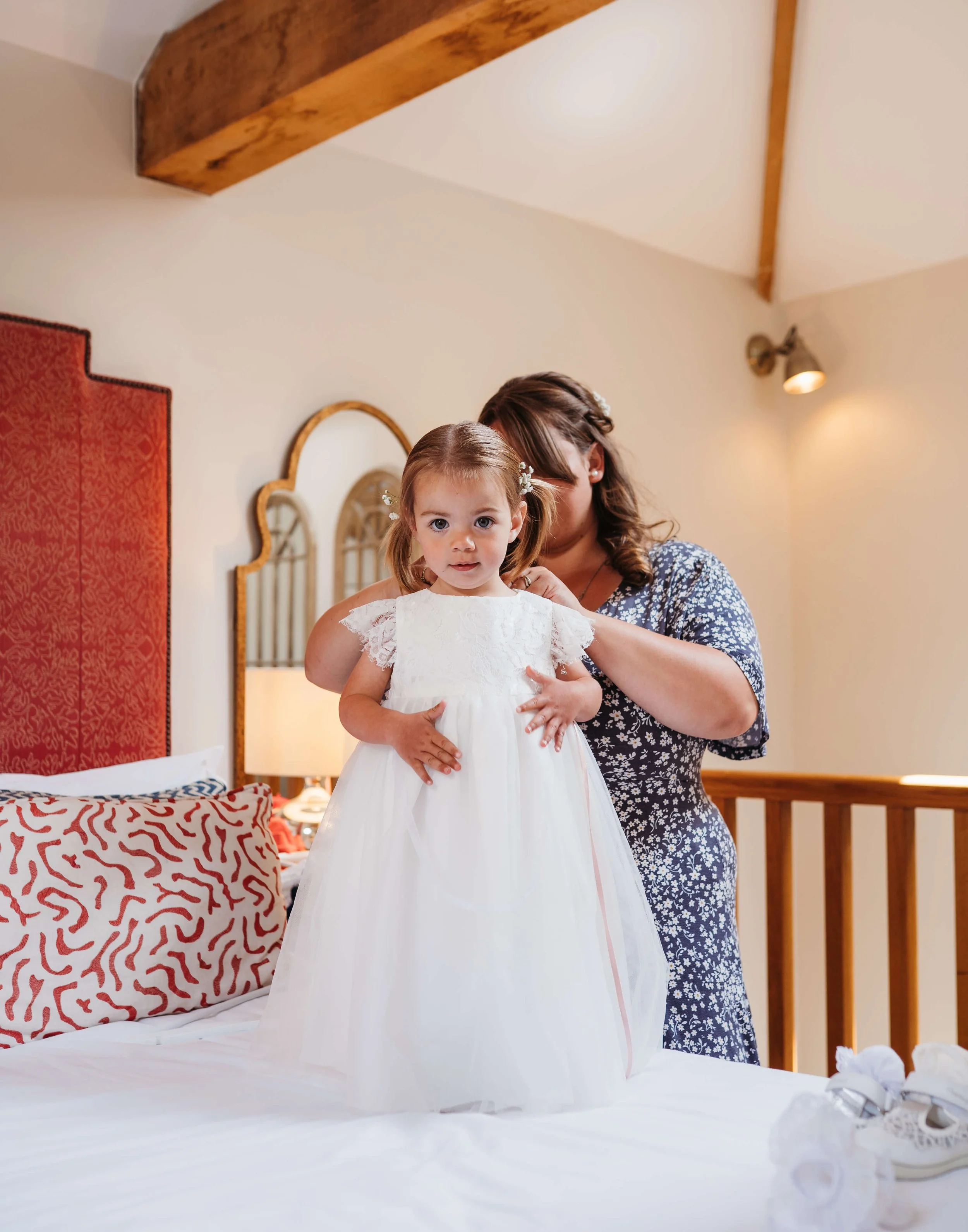 A young girl in a white dress standing on a bed with an adult woman behind her, adjusting her dress, in a bedroom with a red headboard, striped pillow, and wooden decor.