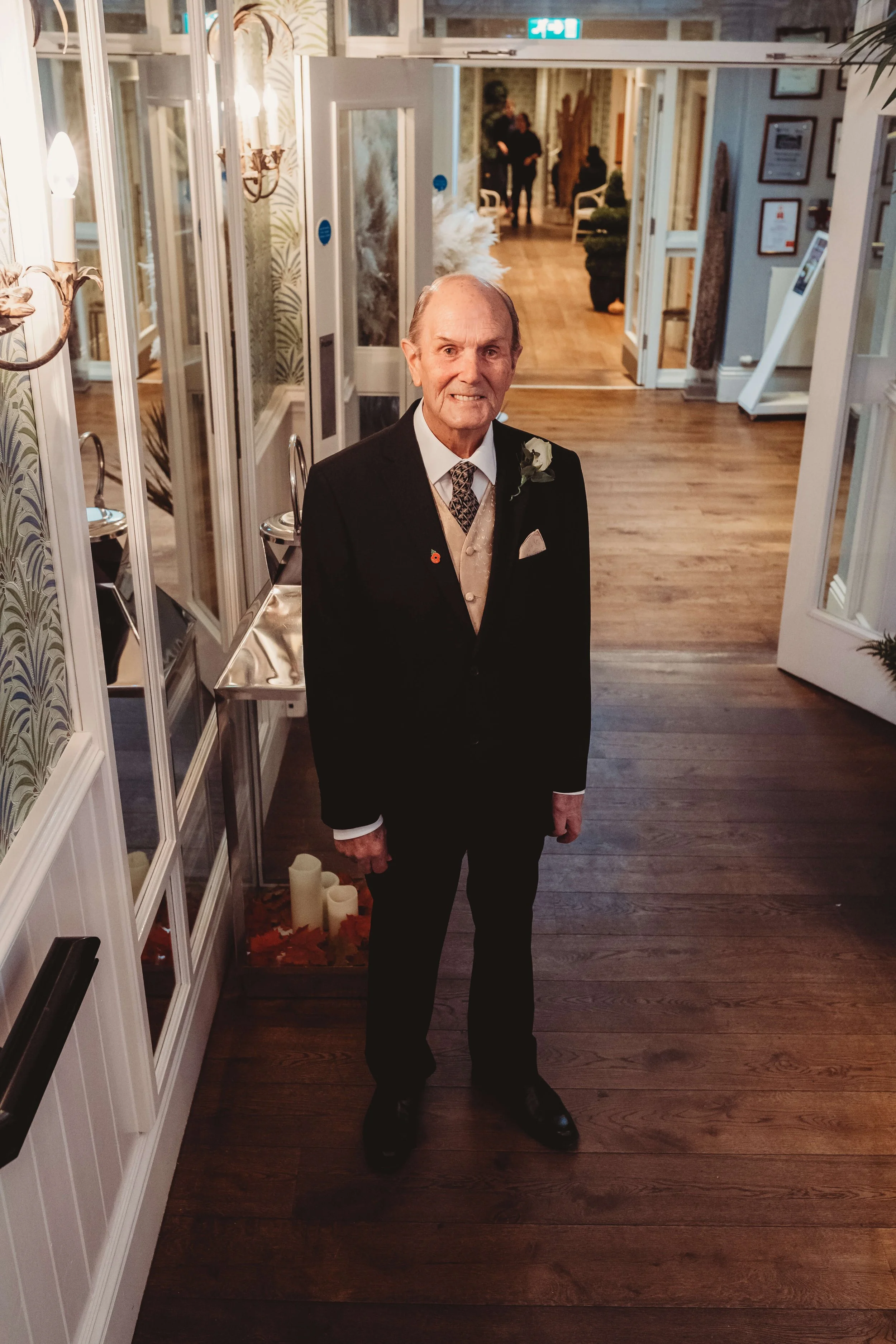An elderly man dressed in a black suit with a white shirt, beige vest, and patterned tie standing inside a decorated venue, possibly a wedding or celebration, with wooden flooring and ornate wall decorations.
