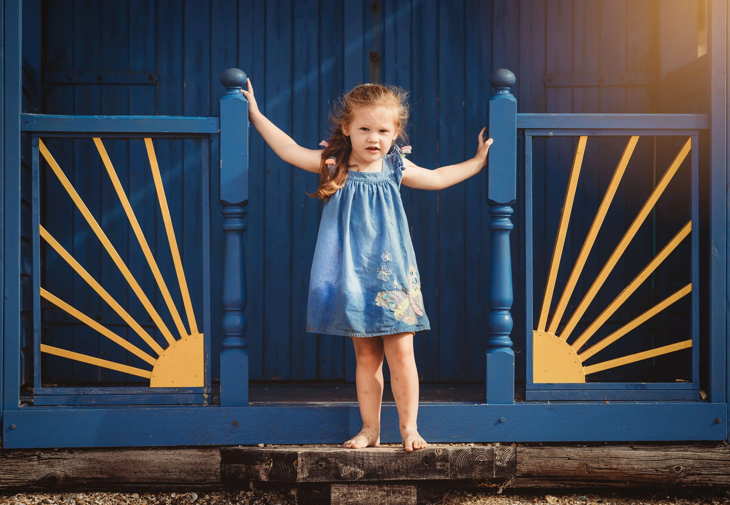 A young girl with curly red hair, wearing a blue dress with butterfly embroidery, standing barefoot on a wooden platform in front of a blue gate with yellow sunburst designs.