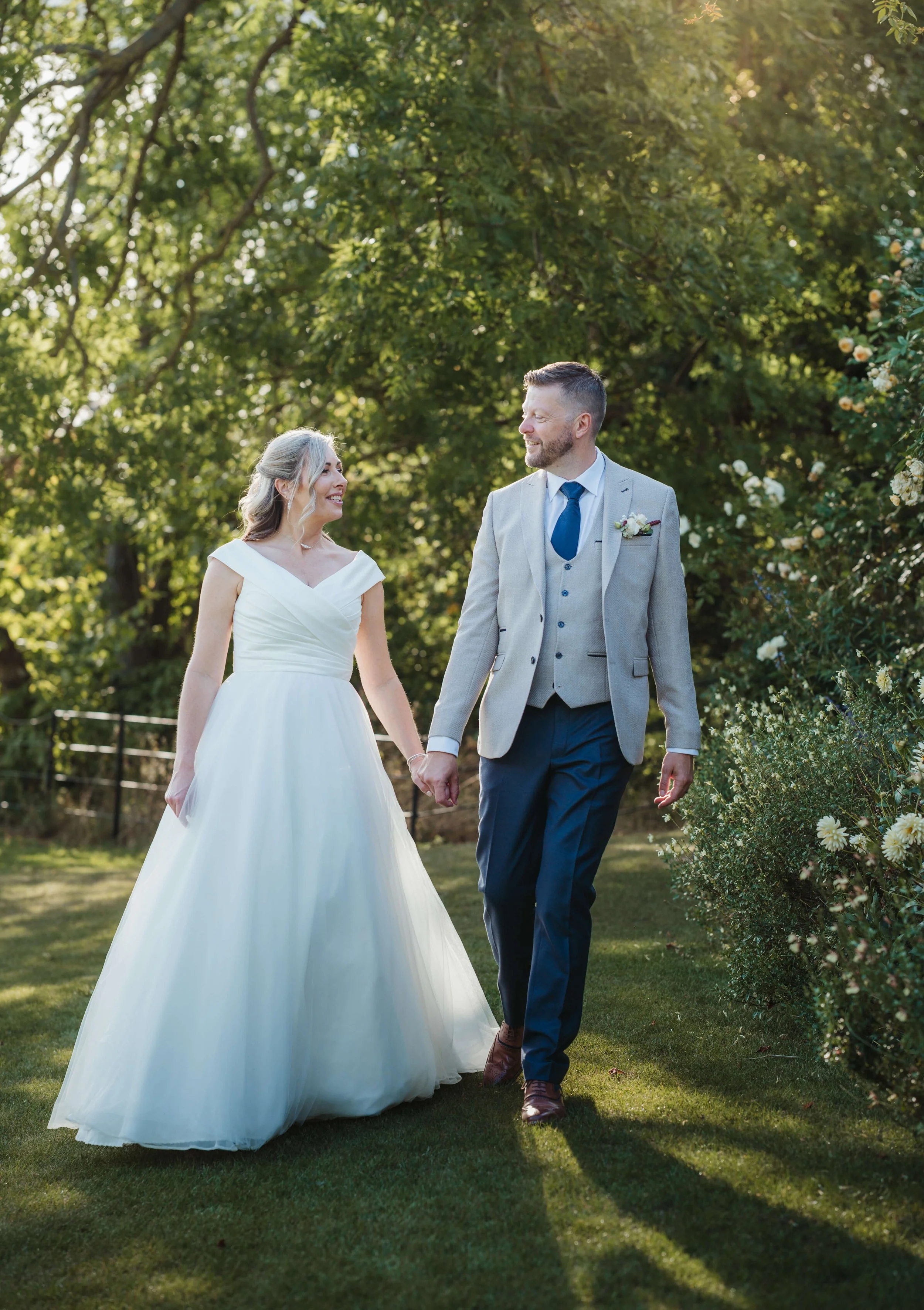 A bride and groom walking hand in hand outdoors during their wedding, surrounded by greenery and sunlight.