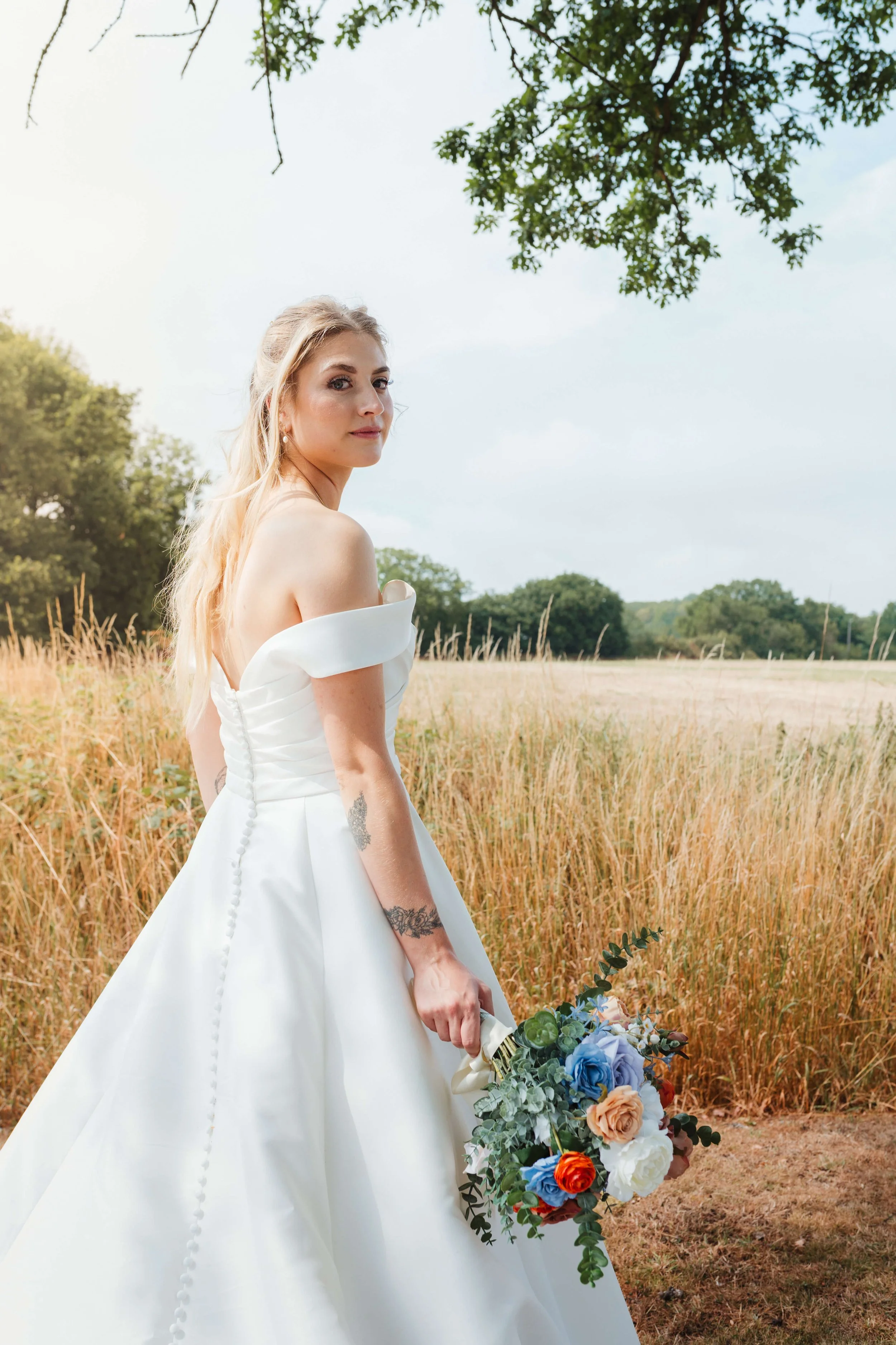 A woman in a white wedding dress holding a colorful bouquet of flowers standing in a grassy field with trees in the background.
