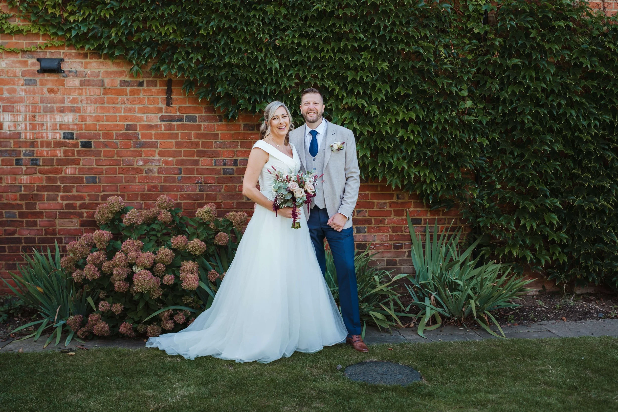 A bride and groom standing outdoors in front of a brick wall covered with green ivy and surrounded by flowers. The bride is wearing a white wedding dress and holding a bouquet, and the groom is in a light gray suit with a blue tie.