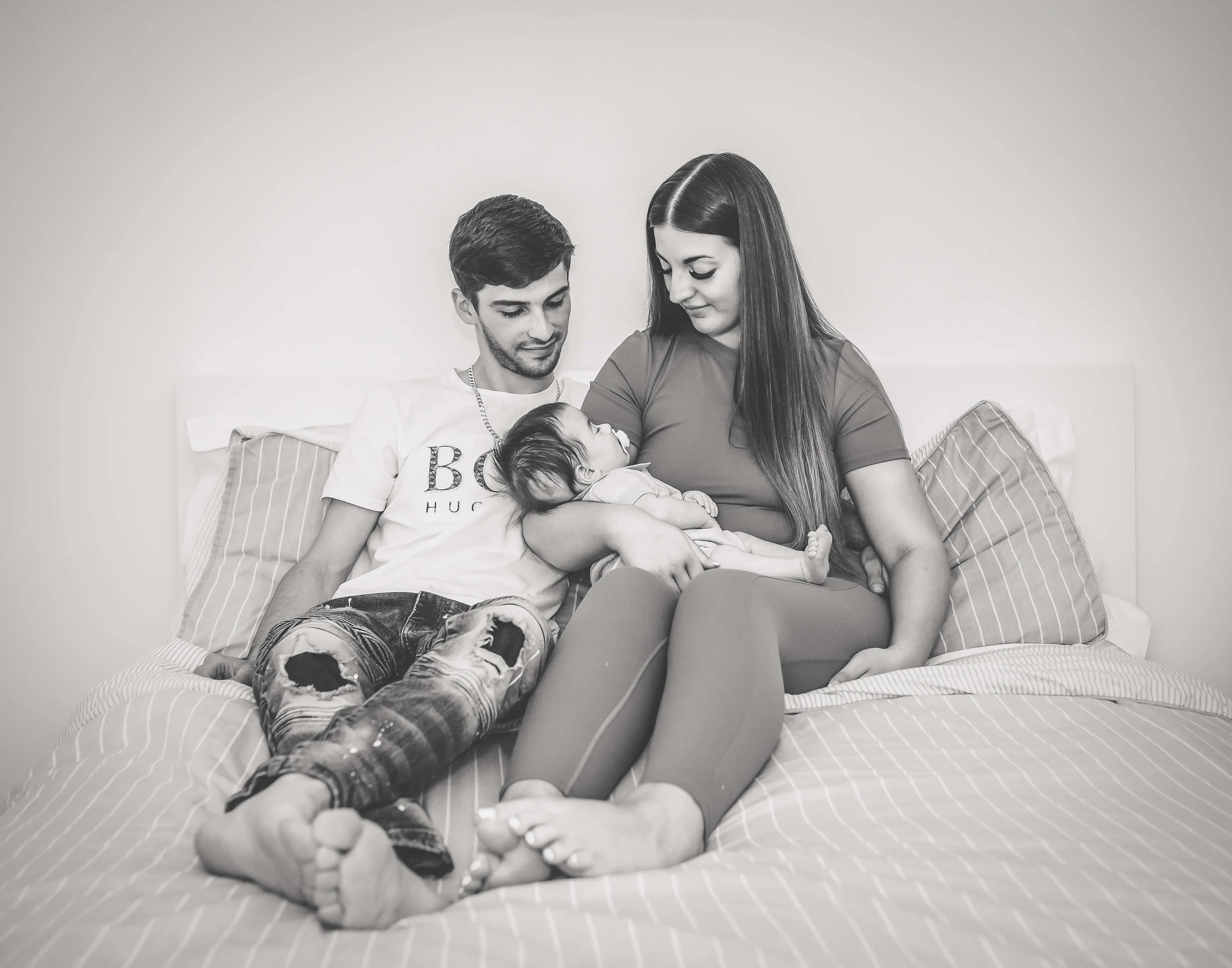 A family of three sitting on a bed with their newborn baby, with the parents looking lovingly at the baby.