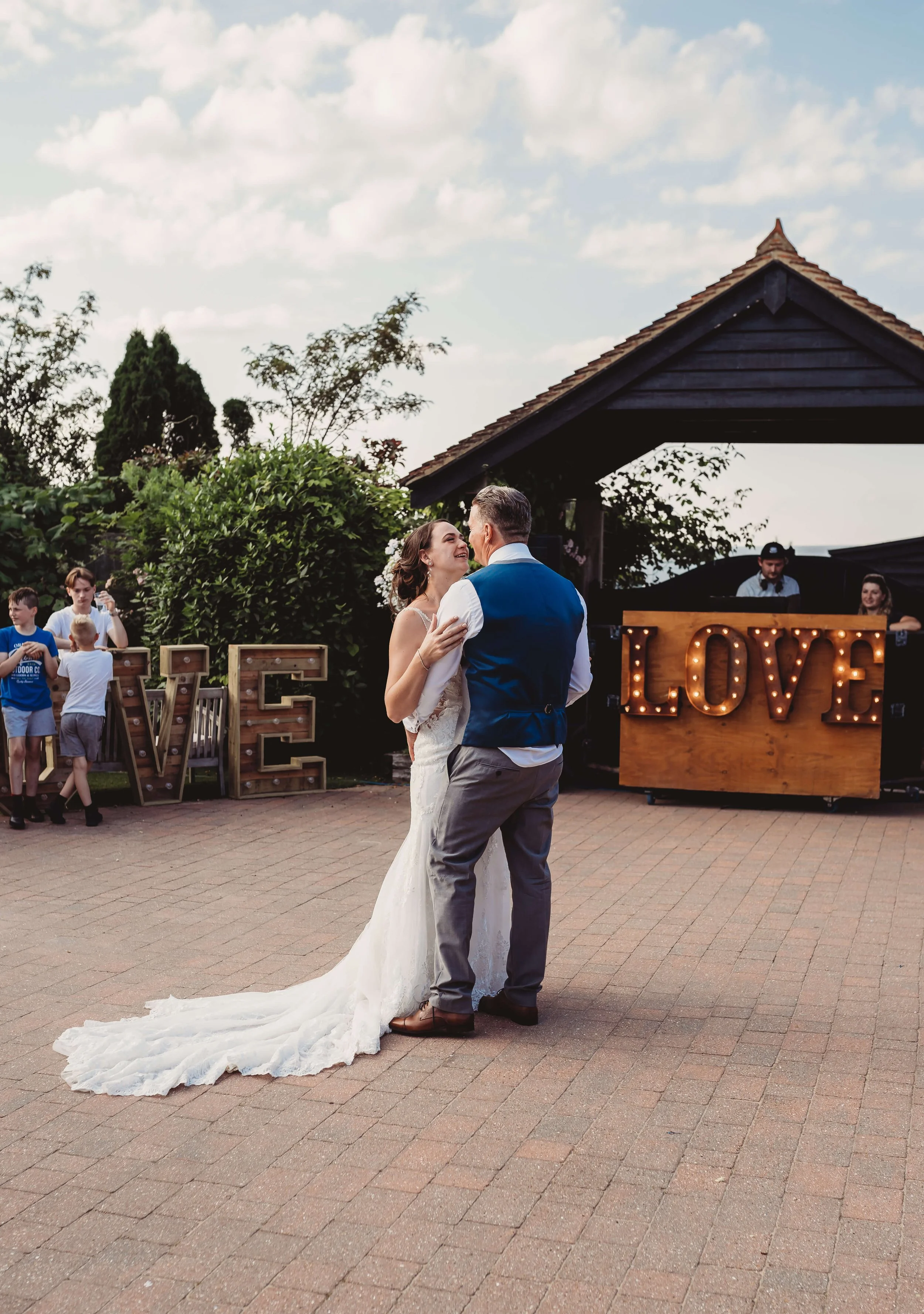 Bride and groom dancing outdoors at a wedding reception, surrounded by friends and decorated with large illuminated love sign.