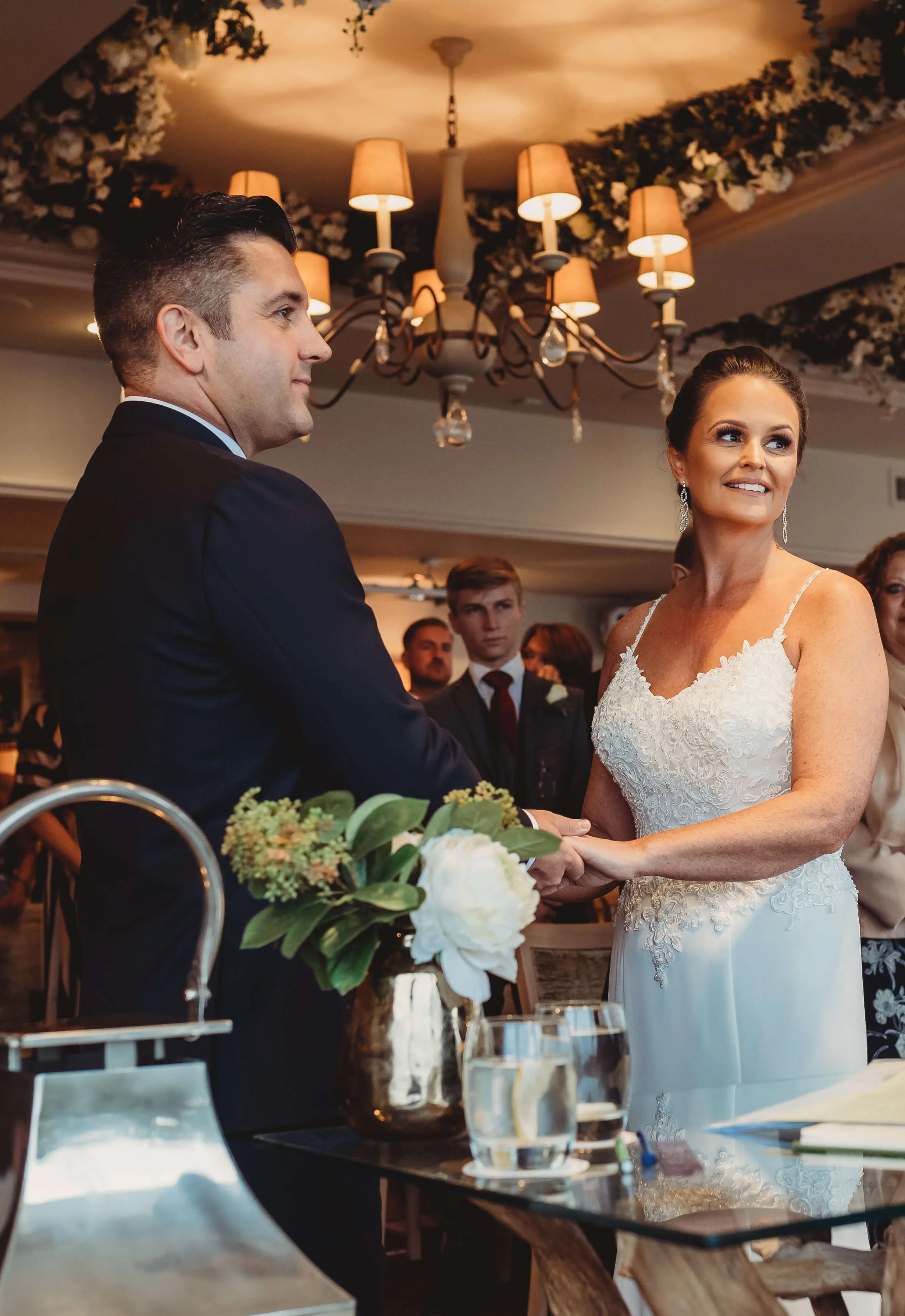 A bride and groom holding hands during their wedding ceremony, with a floral arrangement, glasses of water, and a chandelier overhead in a decorated indoor venue.