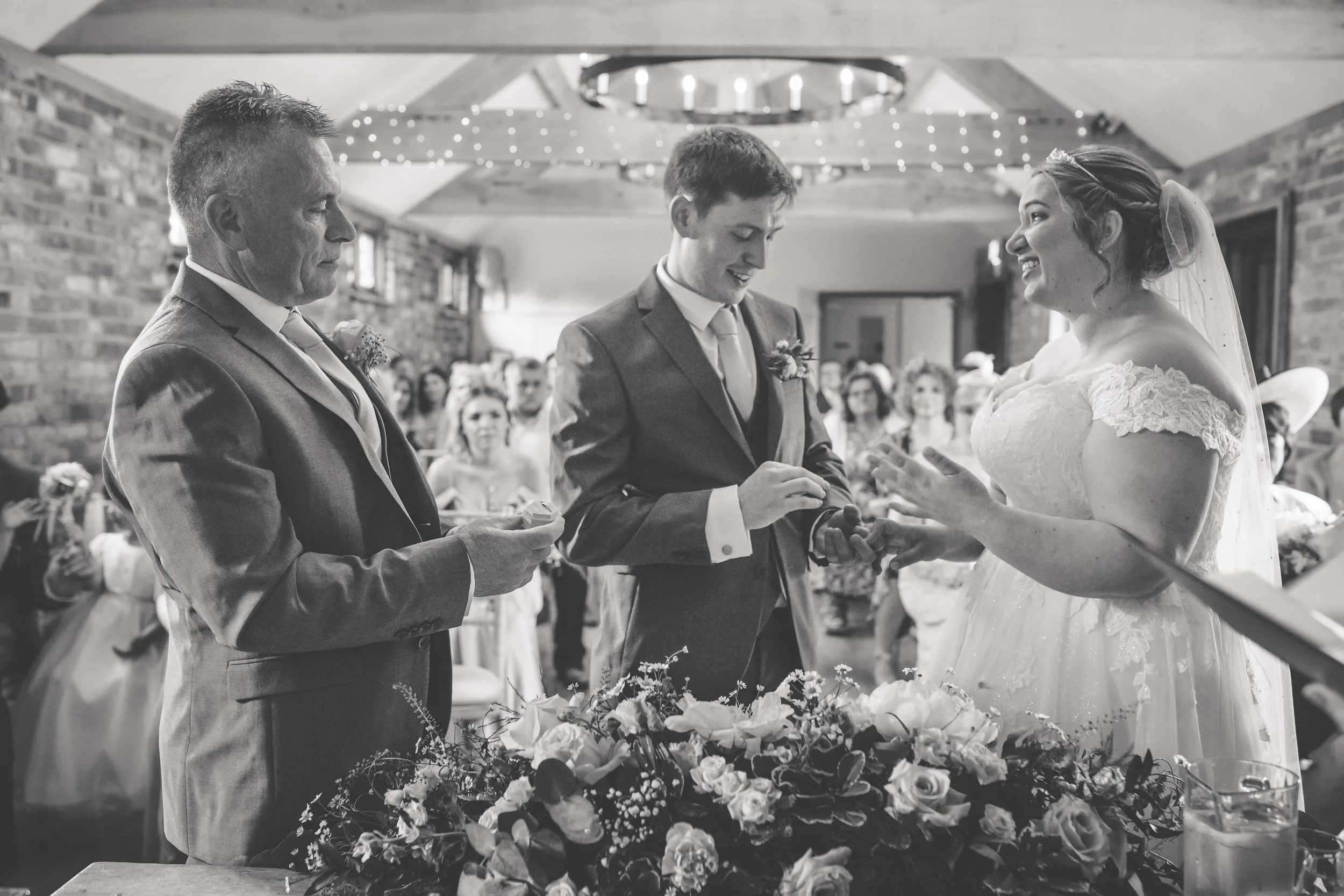 A black and white photo of a wedding ceremony with a bride, groom, and officiant standing in front of guests, surrounded by floral decorations.
