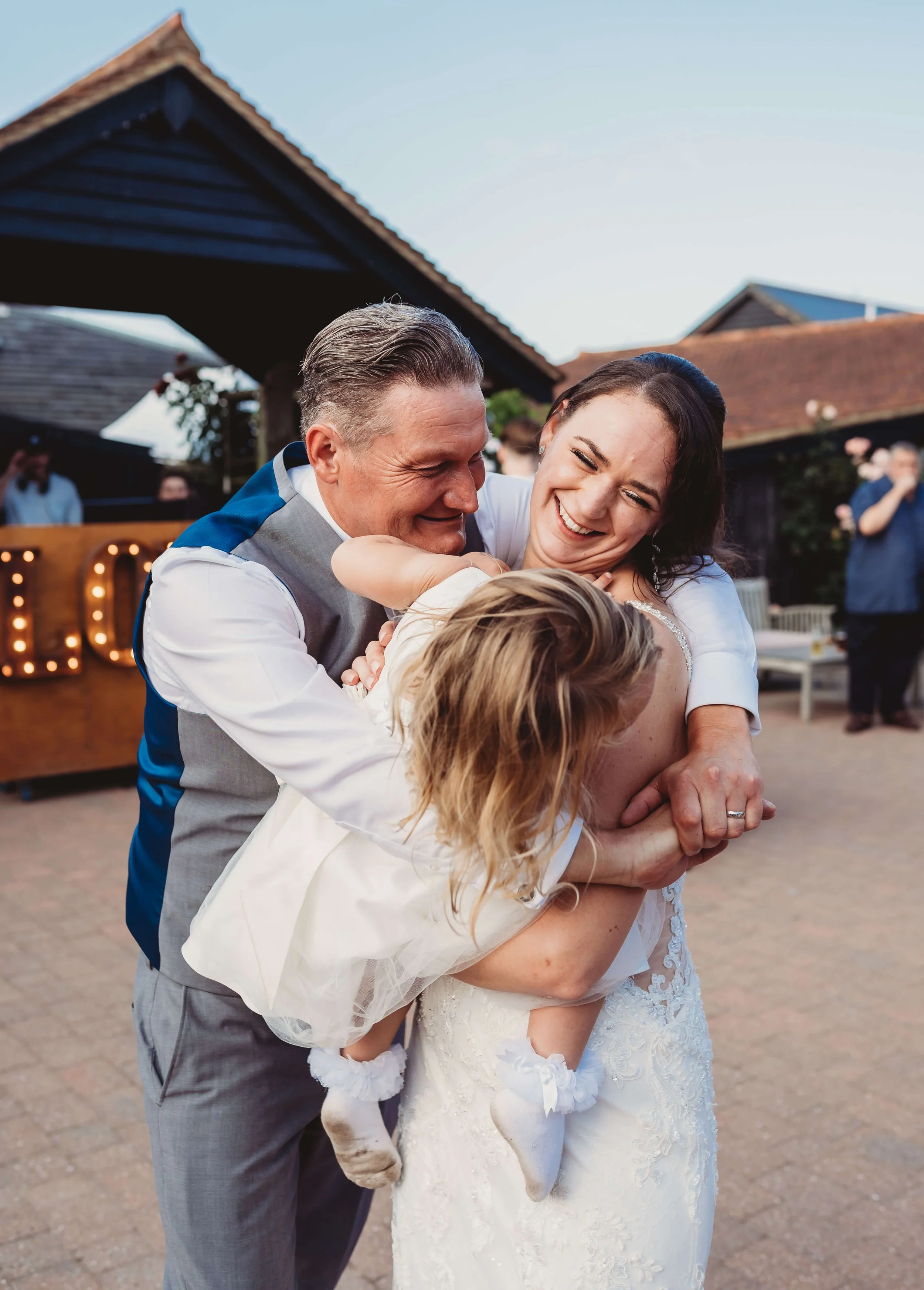 People celebrating at a wedding outdoors. A man and woman are holding a young girl in a dress, smiling and enjoying the moment.