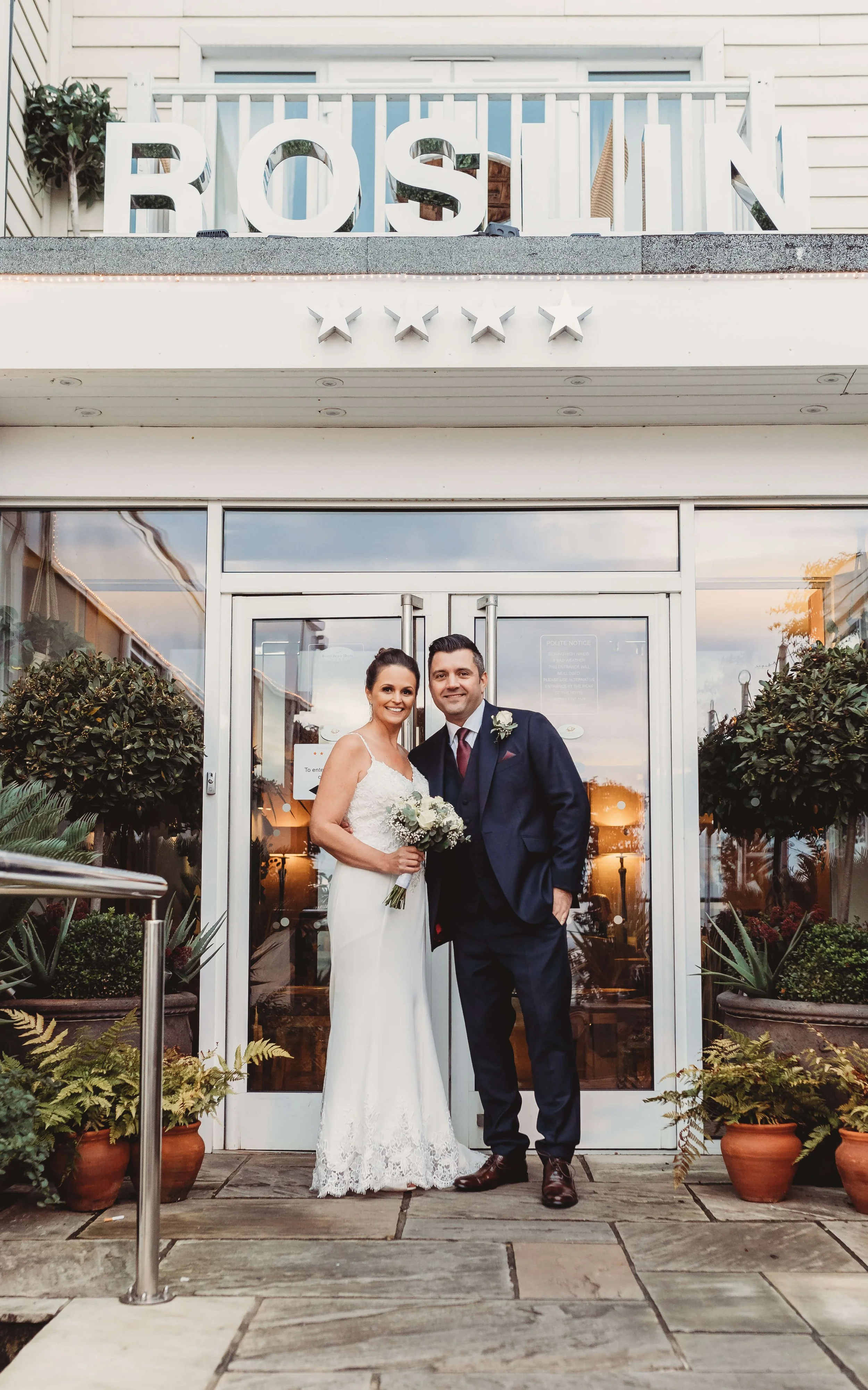 A newlywed couple standing in front of a restaurant with the sign 'ROSTLIN' and four stars, the bride holding a bouquet of white flowers and the groom in a navy suit with a boutonniere, outside on a stone patio with potted plants.