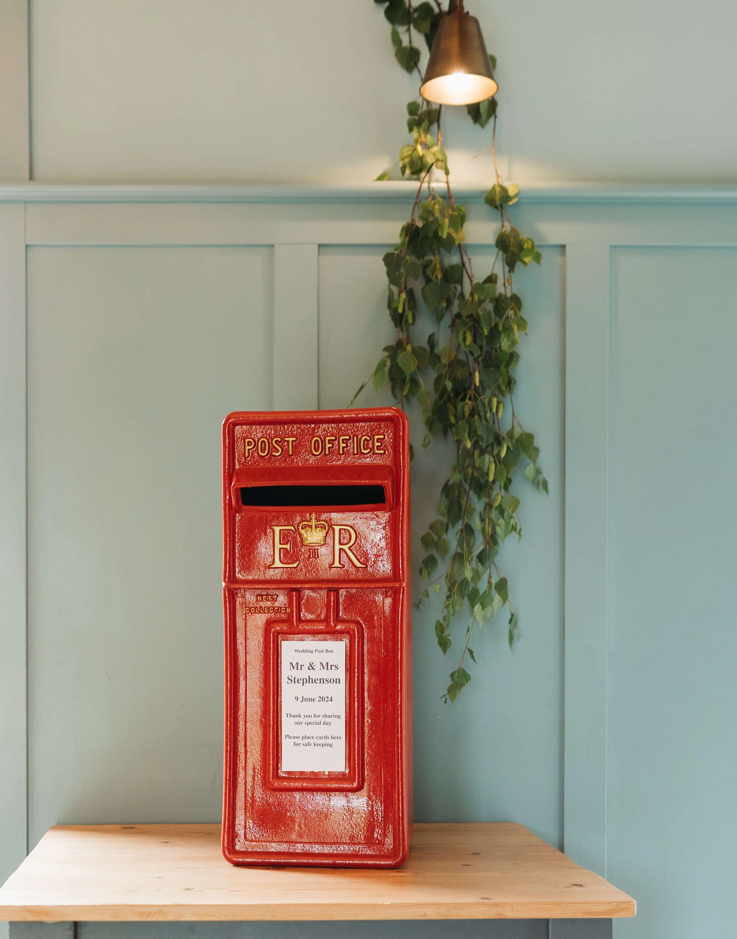 A red British post box with a note attached, placed on a wooden table against a light blue wall with a hanging plant and a wall-mounted lamp.
