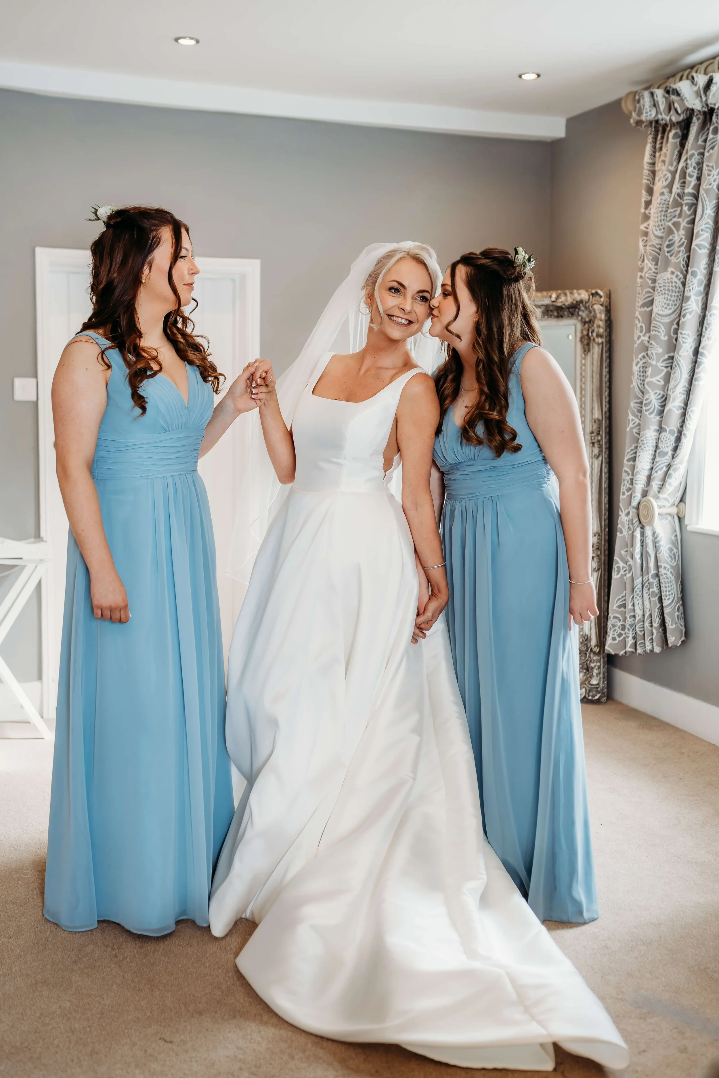 A bride in a white wedding dress is holding hands with two women in blue bridesmaid dresses, standing in a room with gray walls and curtains, sharing a moment of joy.