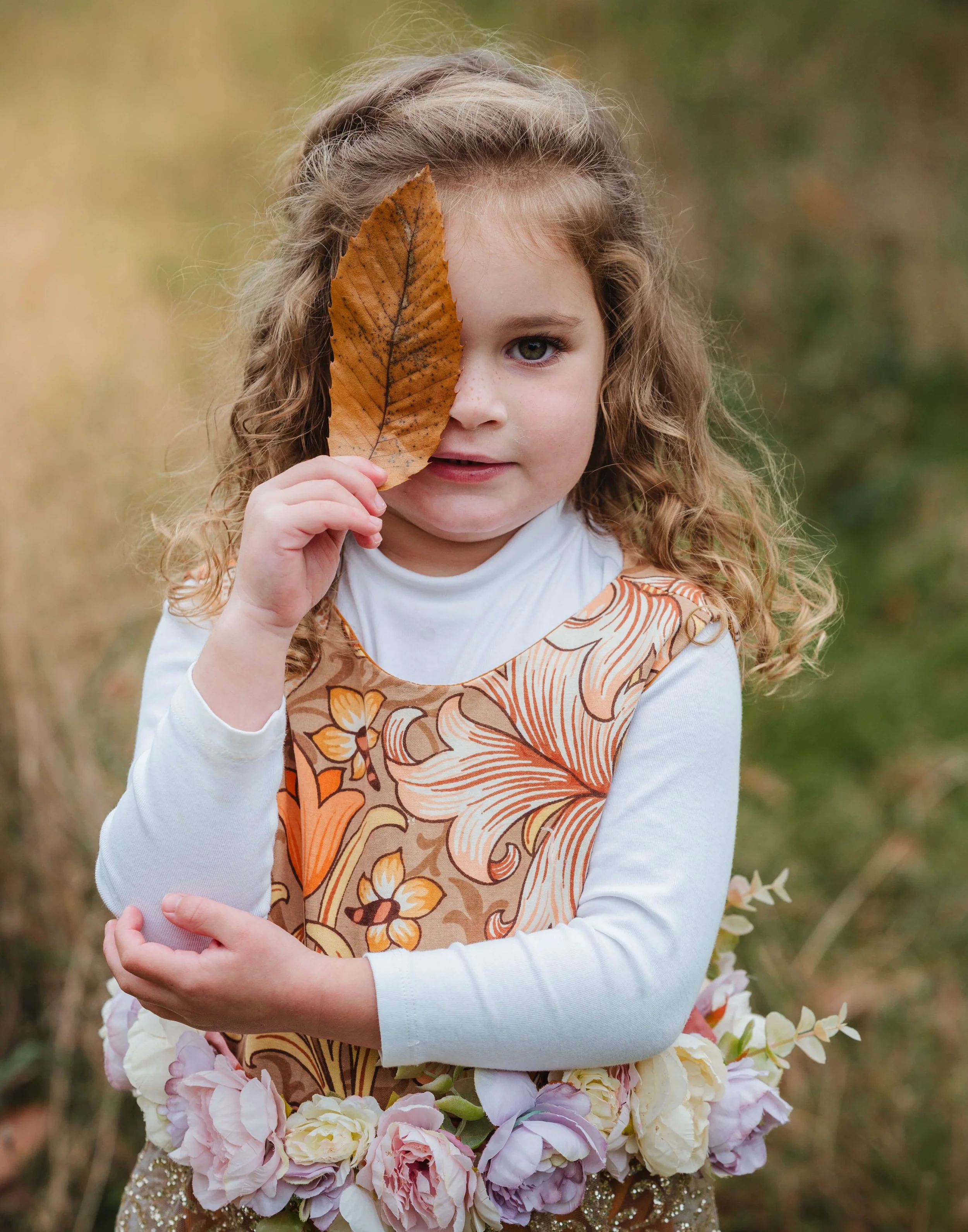 A young girl with curly hair holds a large autumn leaf in front of her face, partially covering her left eye, while standing outdoors surrounded by fall foliage.
