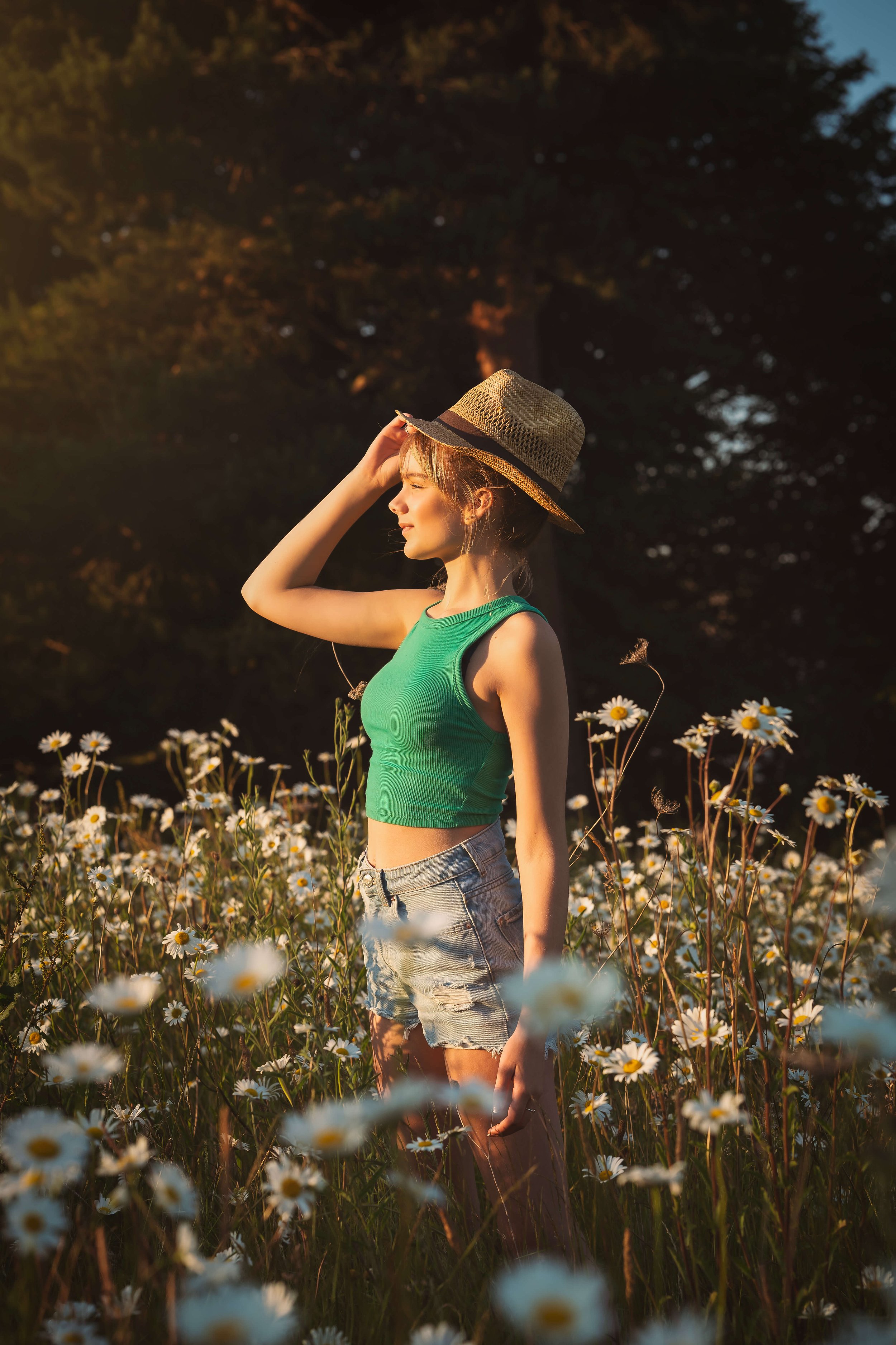 A young woman in a green tank top, denim shorts, and a straw hat standing in a field of daisies during sunset.