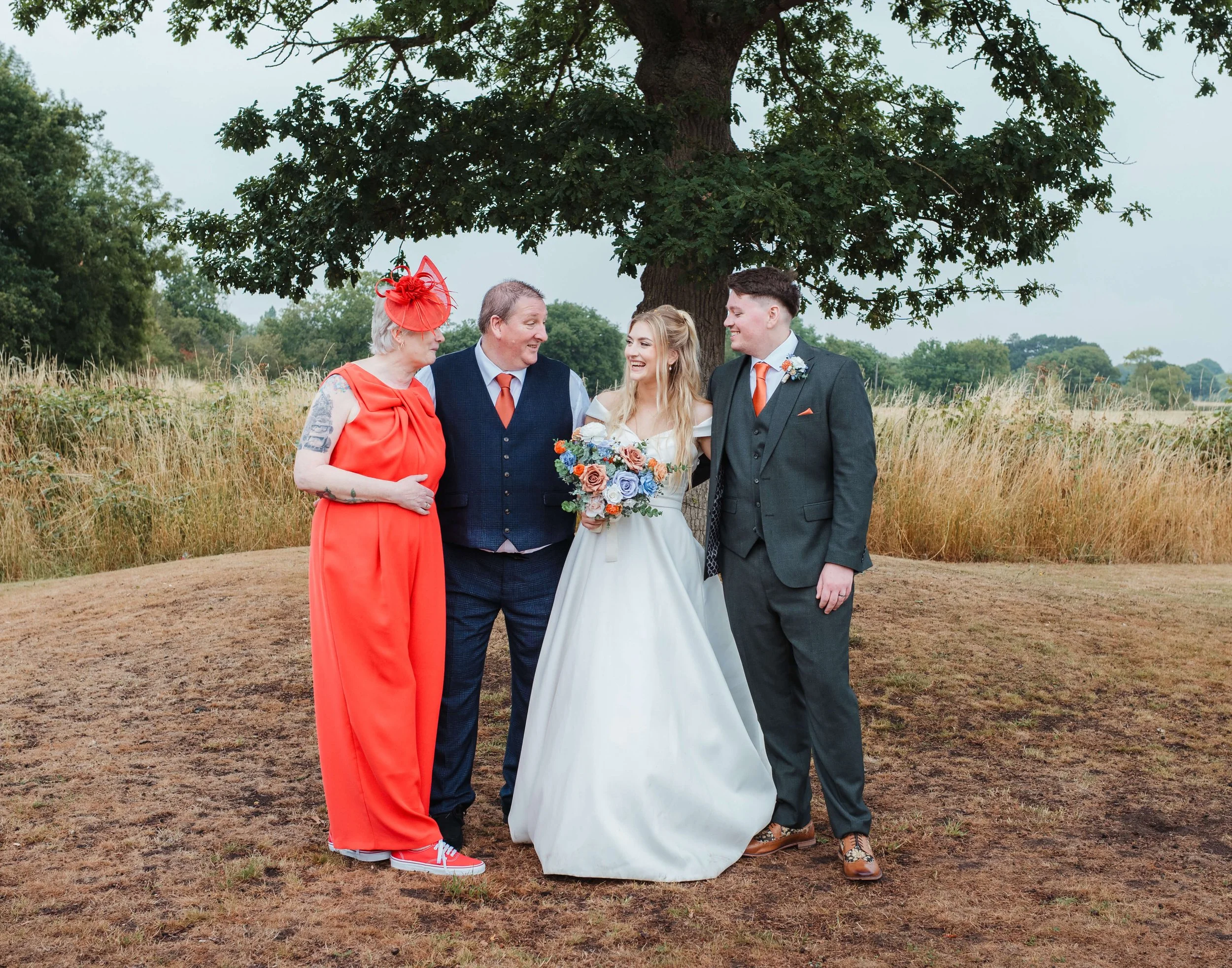 A group of four people, including a bride in a white wedding dress holding a colorful bouquet, standing outdoors under a large tree. They are smiling and talking, dressed in formal and colorful attire.