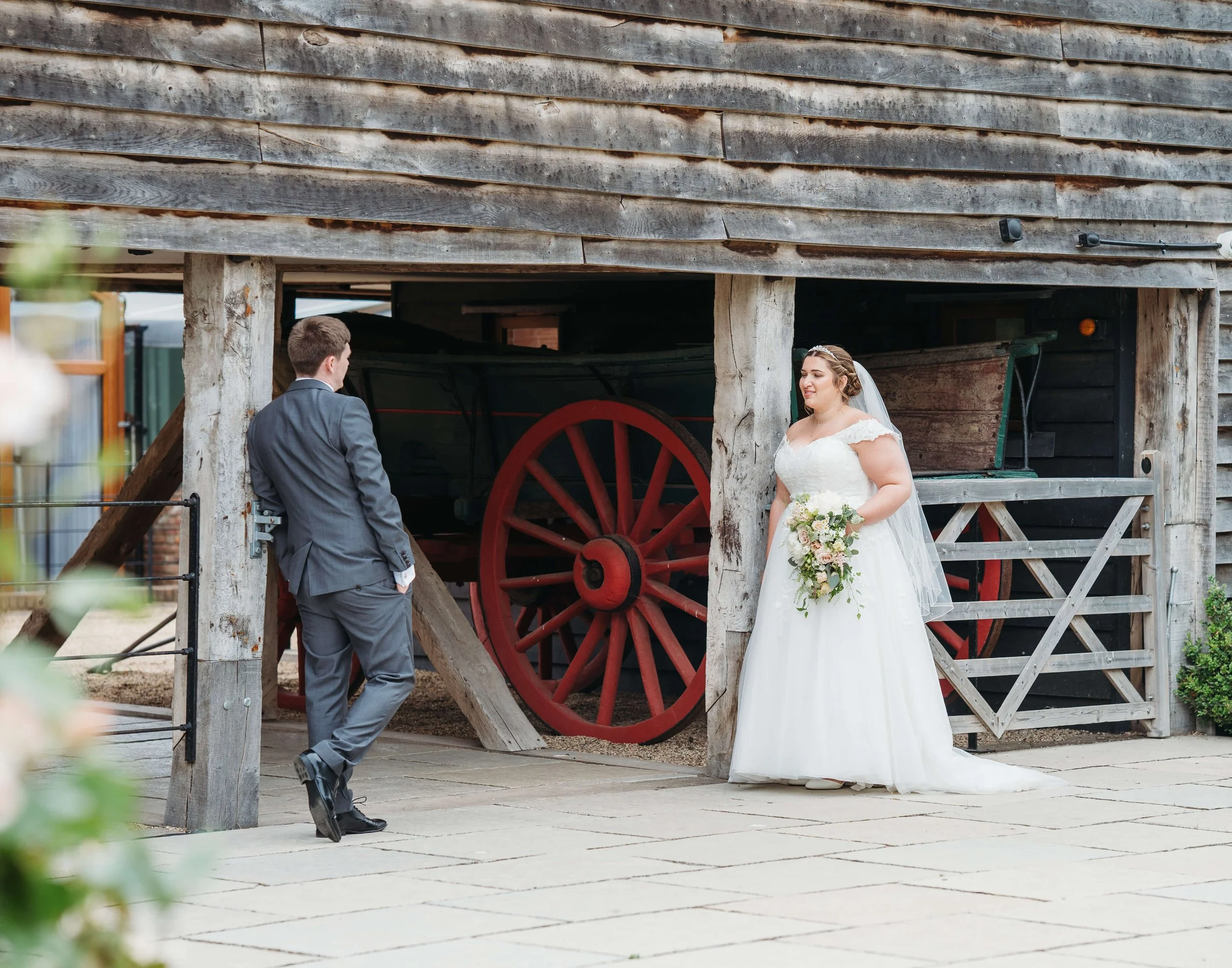 A bride and groom standing outdoors in a rustic setting, with the groom in a gray suit and the bride holding a bouquet, near an old wooden structure with a vintage wagon.