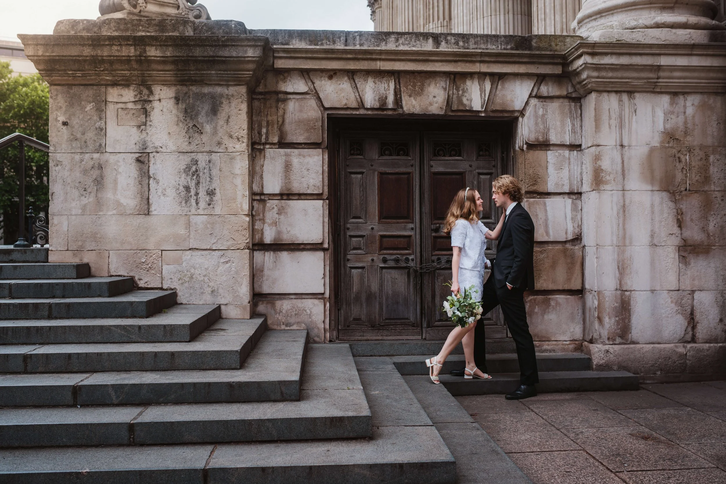 A bride and groom standing face to face in front of a large wooden door, with chain locks, on a stone building. The bride wears a white dress and heels, holding a bouquet, and the groom wears a black suit. They are close and looking at each other, wi