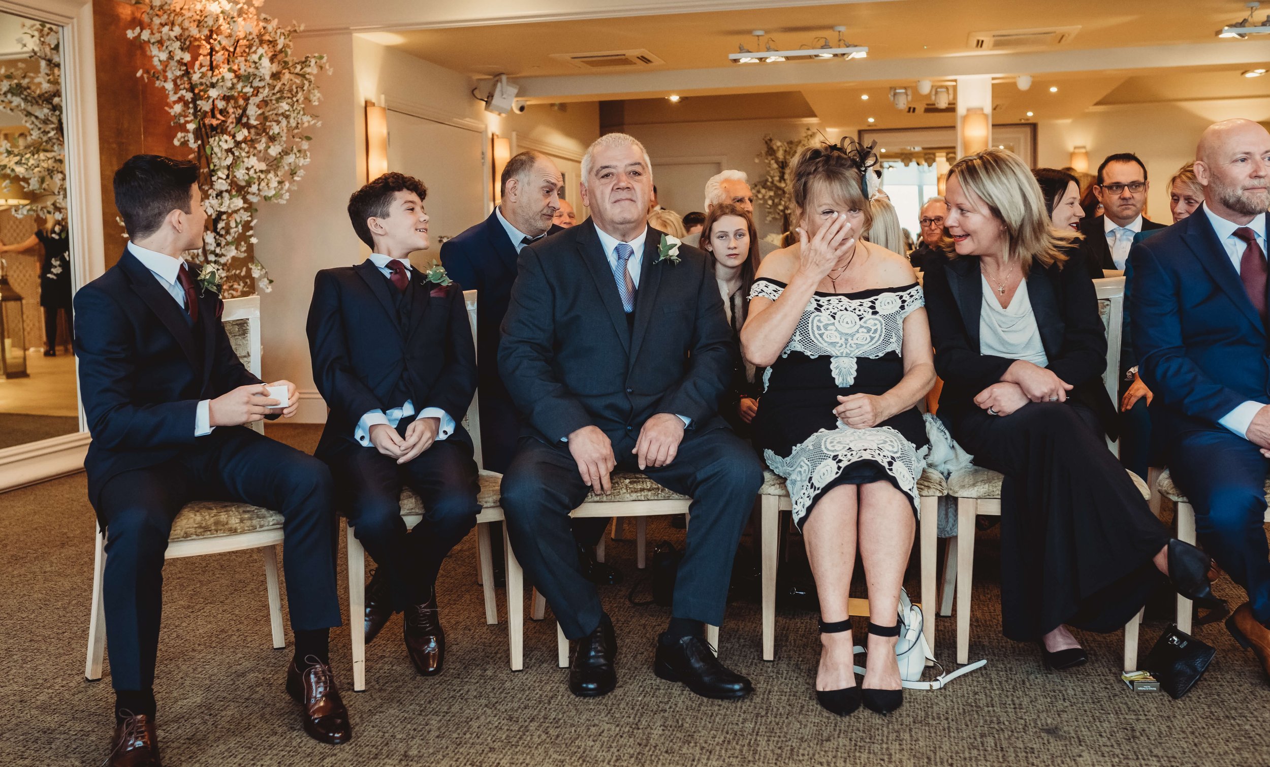 A group of people seated in chairs at a wedding ceremony, with two boys in suits on the left, an older man, woman, and another woman in the middle, dressed in formal attire, and others in the background.