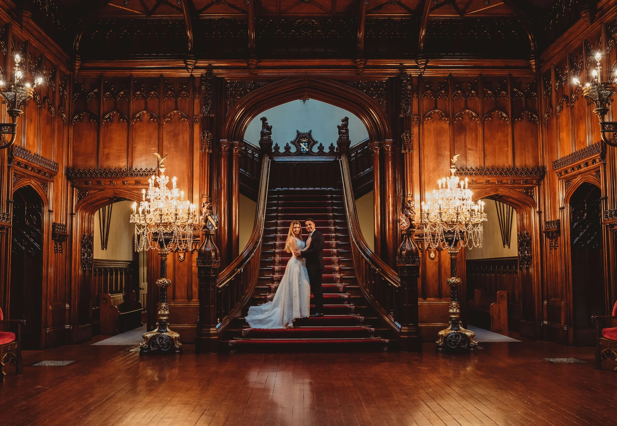 A bride and groom in wedding attire standing on a grand staircase inside a wood-paneled mansion with ornate chandeliers.